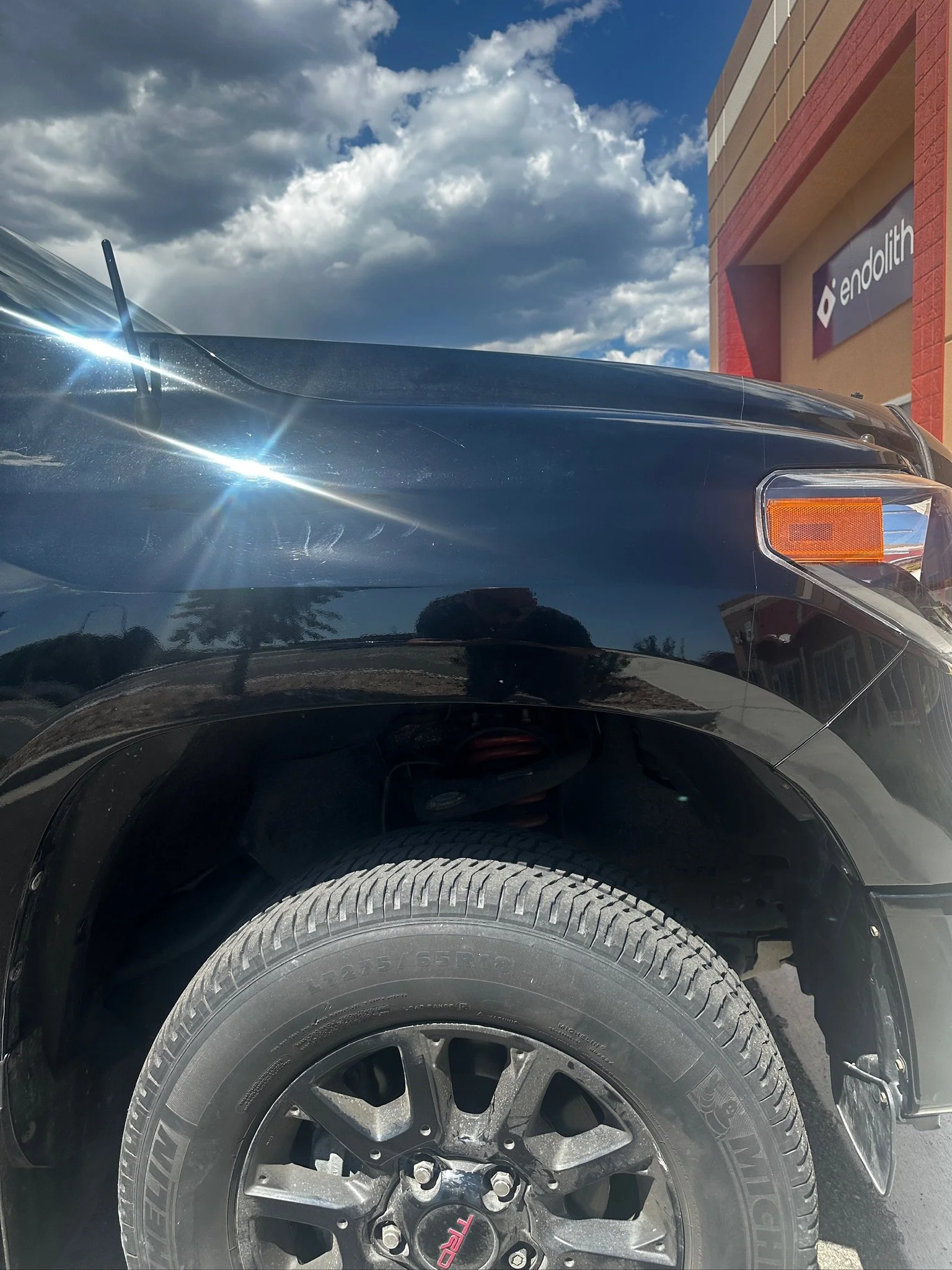 Close-up of a black car's front wheel, tire, and part of the front fender, with a building and cloudy sky in the background.