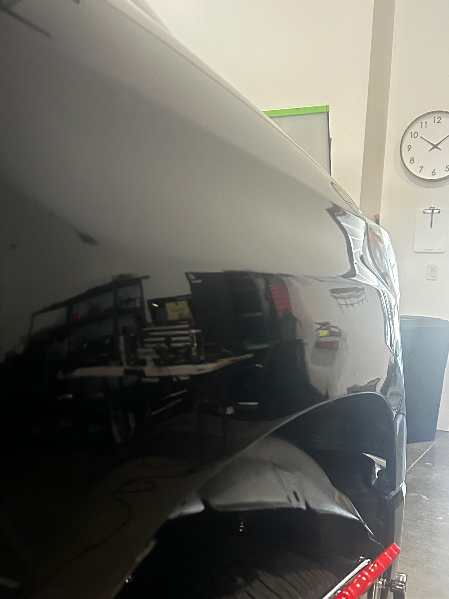 The side of a black car parked in an indoor parking garage. In the background, a white wall with a clock showing 10:08, a partially visible whiteboard with green trim, and a red fire extinguisher are visible.