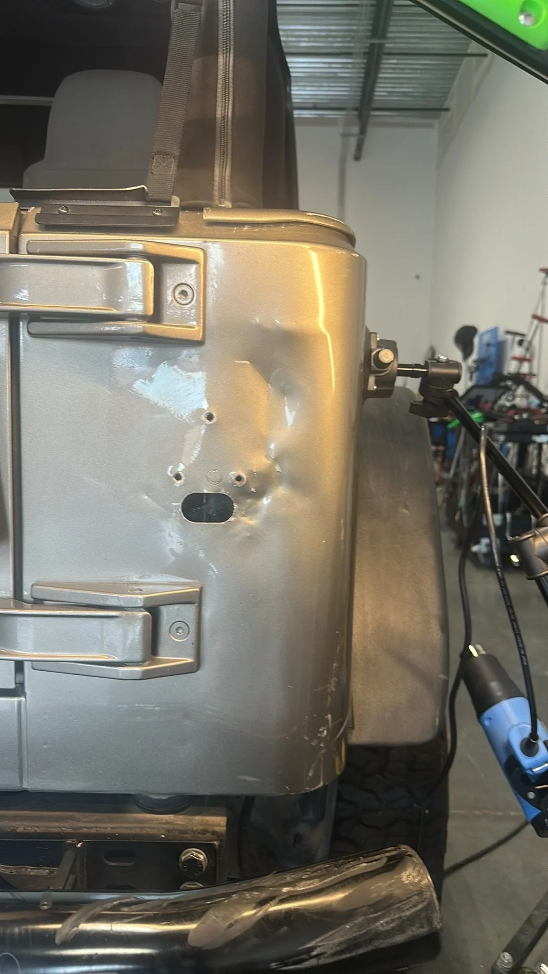 Close-up of a metallic vehicle component, possibly a part of a truck or heavy machinery, with a dent and some scratches. A mounting bracket with a bolt is visible on the right side. In the background, there is a garage or workshop with tools and equi