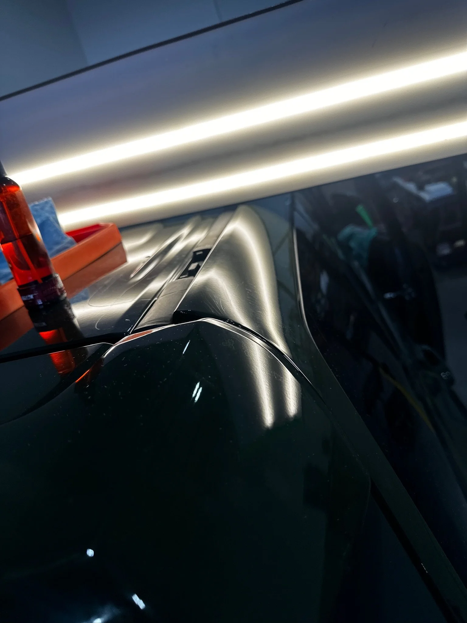 Close-up of a dark-colored car with reflective surface in a garage, with bright overhead lighting, tools, and equipment visible in the background.