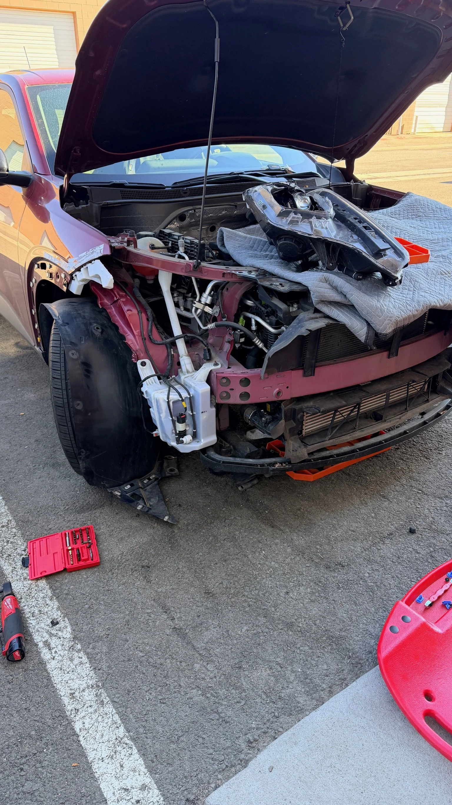 A red car with its front bumper and grille removed, undergoing repair or maintenance, with tools such as a red toolkit, a cordless drill, and a red creeper lying on the ground nearby.