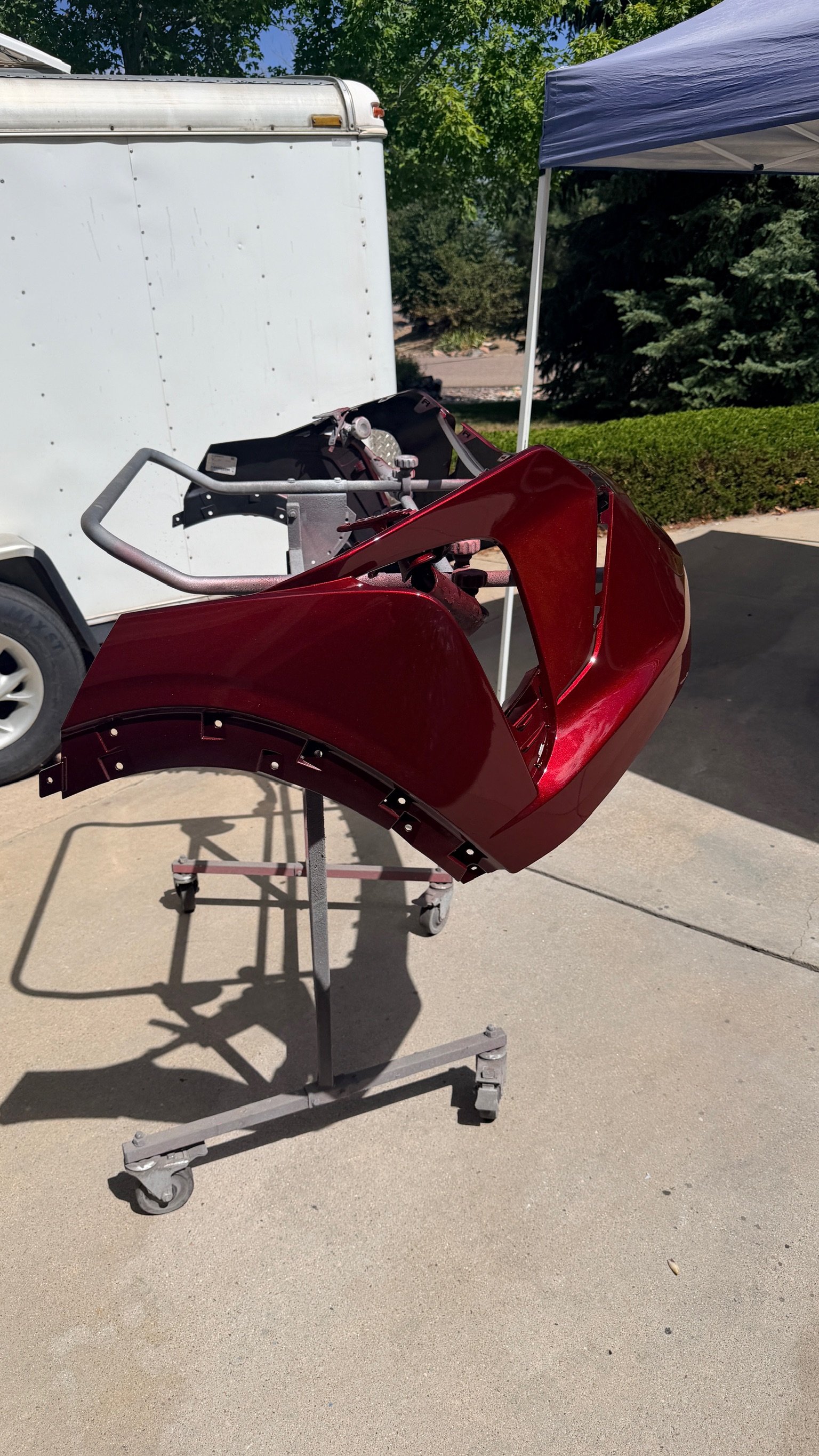 Red motorcycle fairing mounted on a mobile stand outside with a white trailer and green trees in the background.