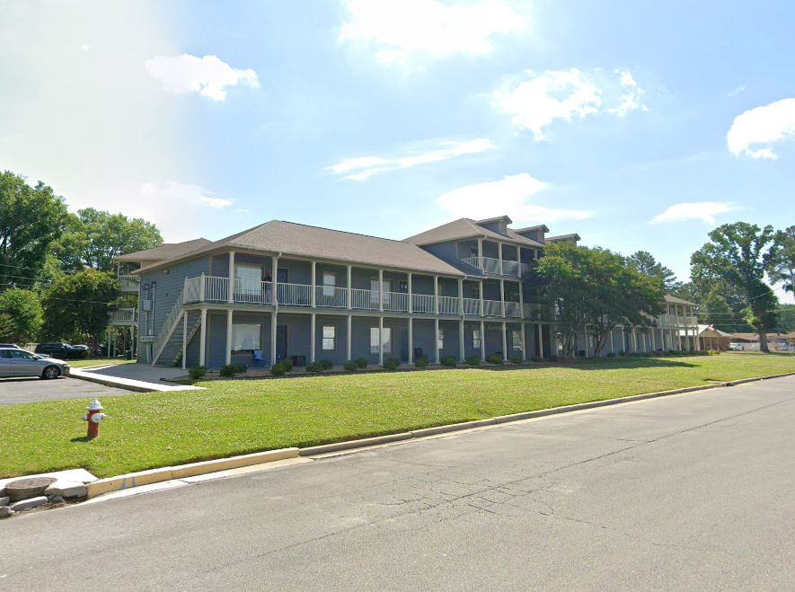 Multi-story residential or apartment building with blue siding, balconies, and stairs outside, set in a grassy area with a sidewalk and parking lot in front, under a partly cloudy sky.