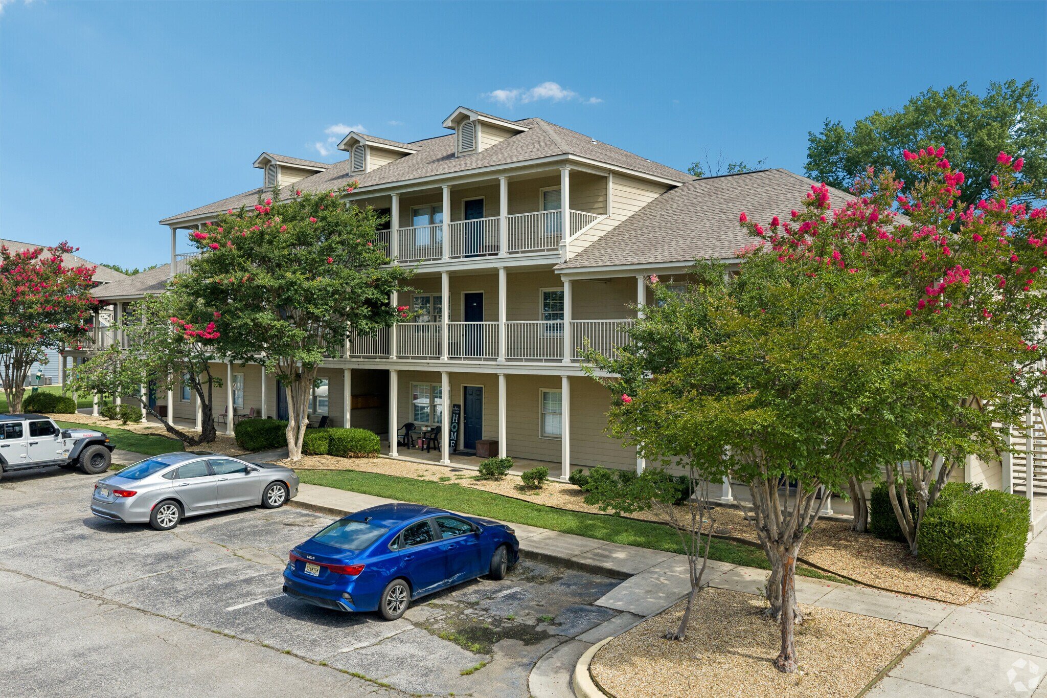 A multi-story residential building with beige siding, white trim, and a gabled roof, surrounded by trees with pink flowers. There is a parking lot with several cars in front of the building.