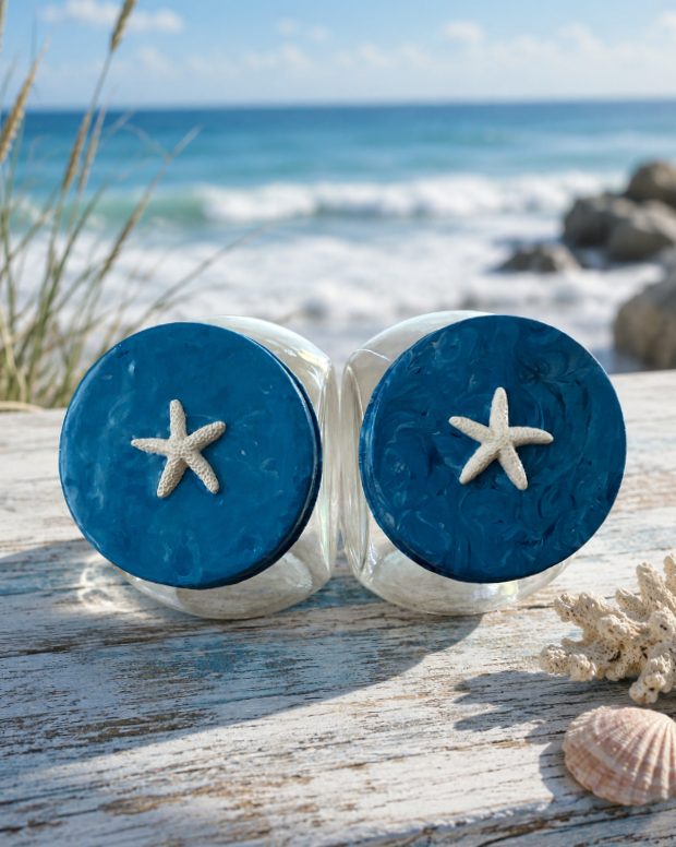 Two glass jars with blue tops and starfish decorations, placed on a weathered wooden surface near the beach, with sea, rocks, and sand in the background.