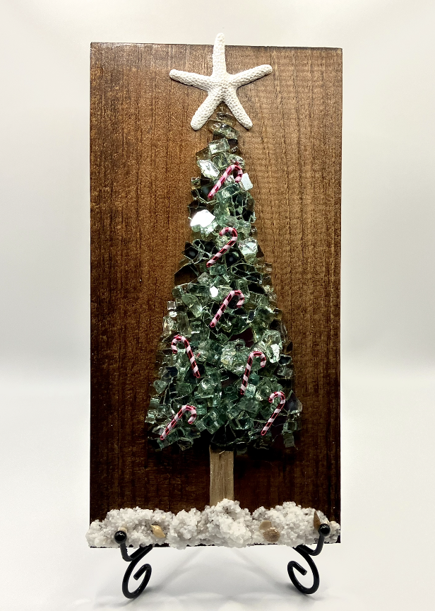 Decorative Christmas tree made of broken glass pieces on a wooden board, topped with a starfish, with candy canes and a snowy base, standing on a black metal stand.