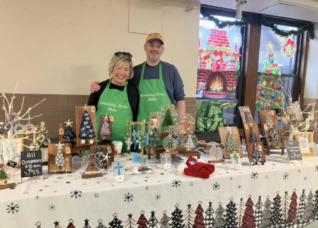 Two people standing behind a table with Christmas ornaments and decorations at a holiday market. They are wearing green aprons and smiling.