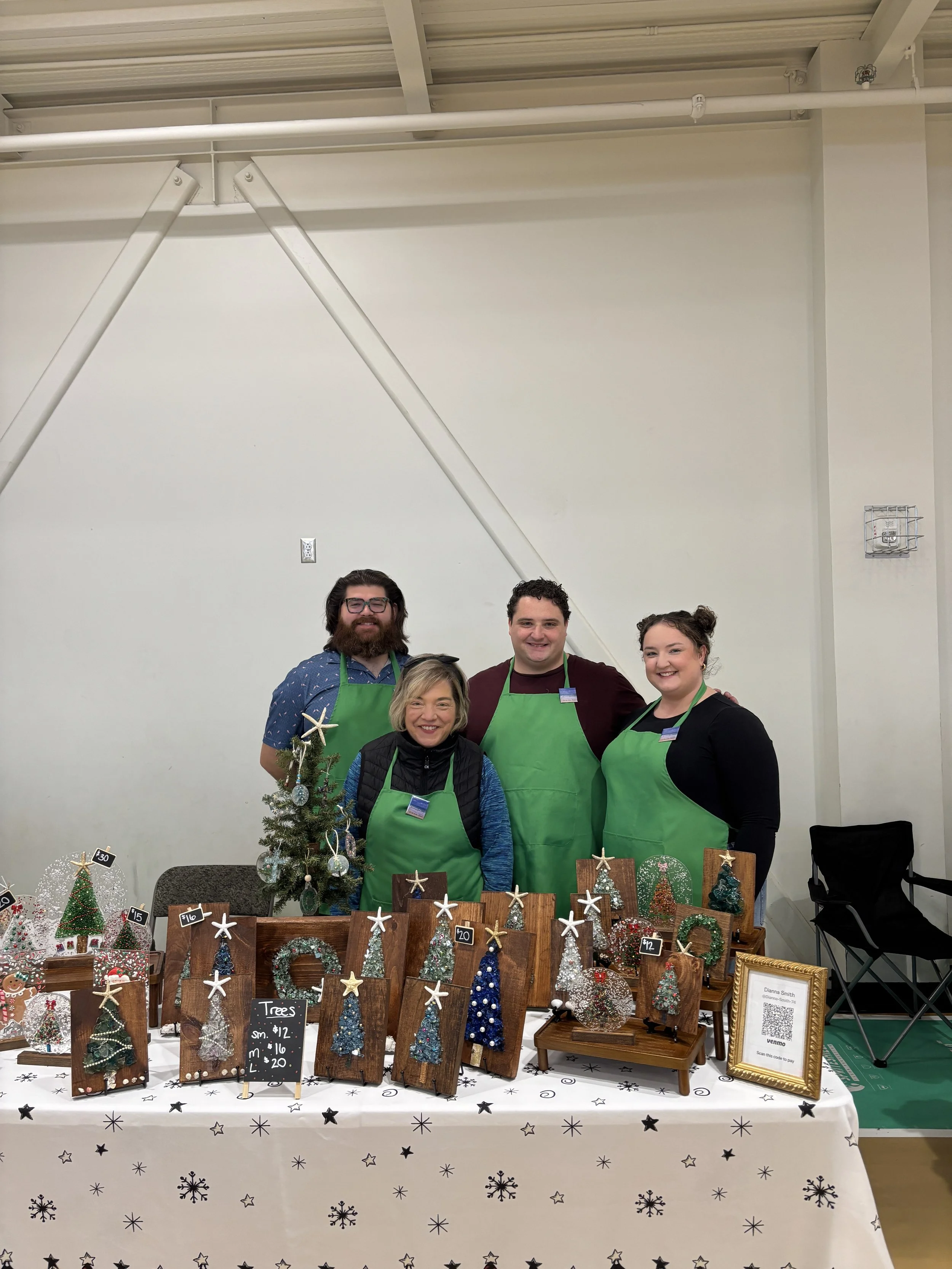 Group of five people standing behind a table with Christmas-themed crafts including small decorated trees, wreaths, and ornaments for sale at a holiday market.