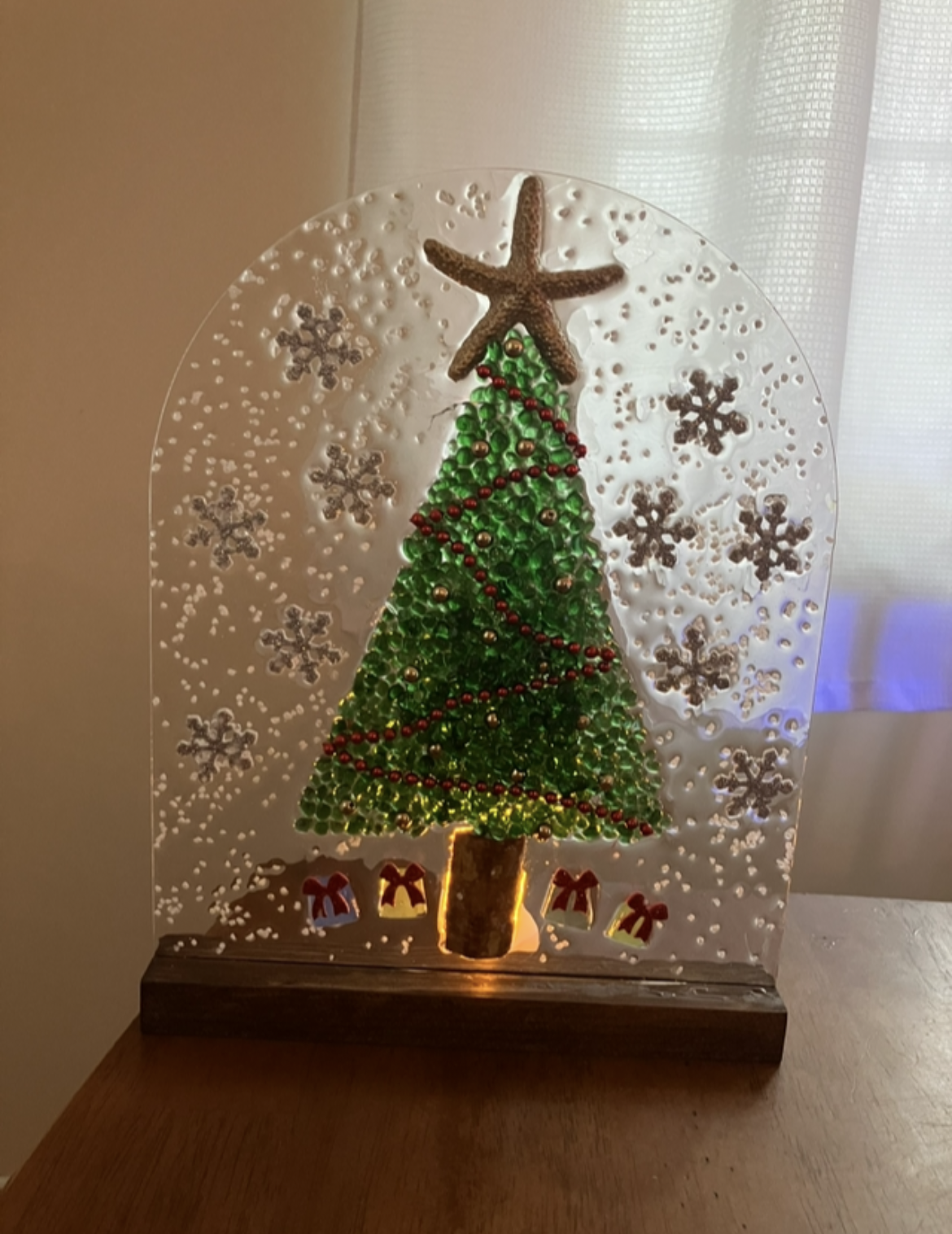 Decorative Christmas display featuring a green Christmas tree with red and gold ornaments, topped with a starfish, and surrounded by snowflakes and gift boxes illuminated from below.