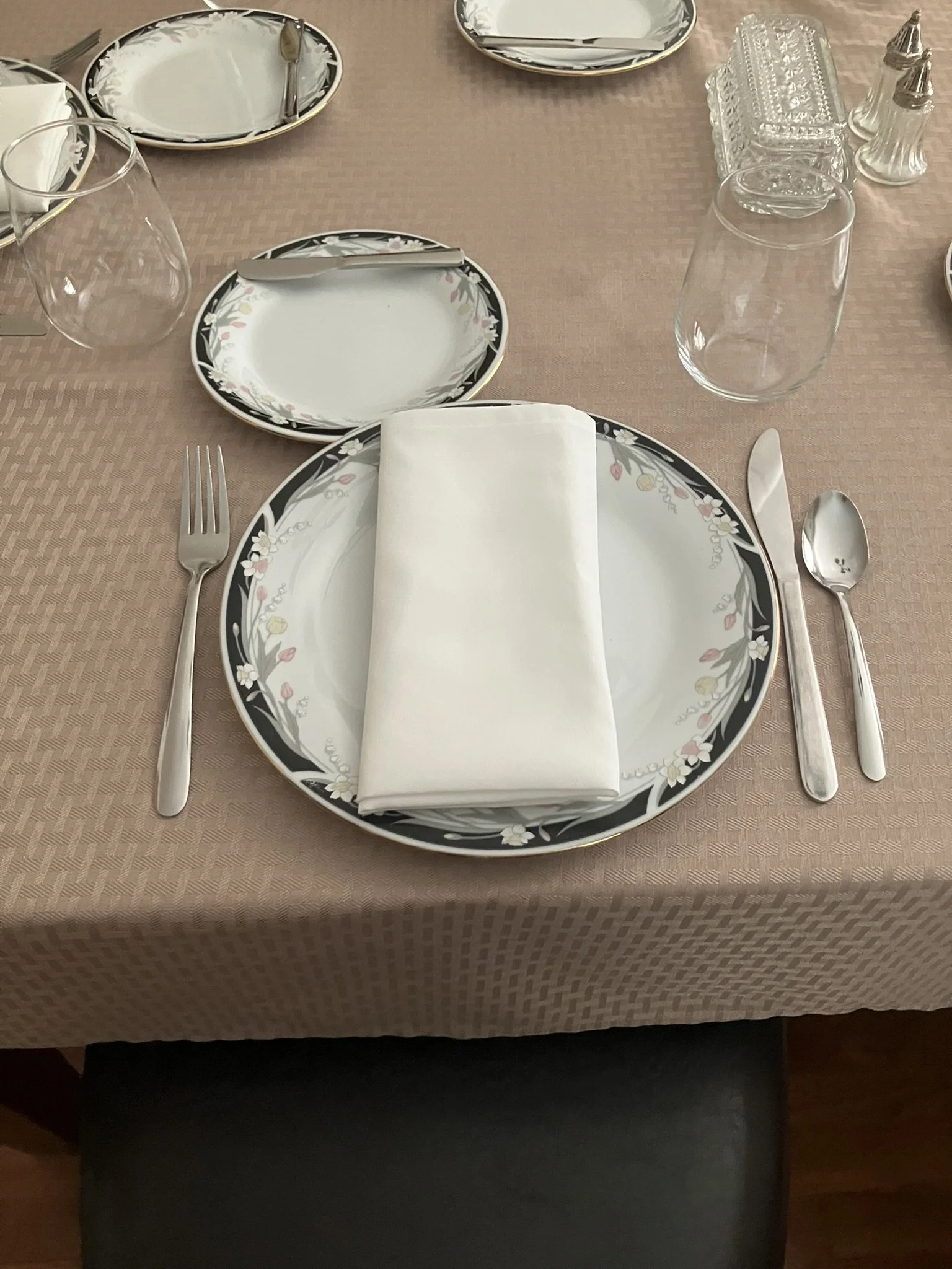 A formal table setting with floral patterned plates, a rolled white linen napkin, silverware, empty wine glasses, and small condiment containers on a beige tablecloth.