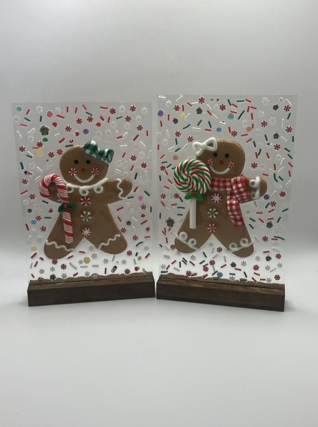 Two gingerbread cookies decorated with icing, each holding a candy cane and a lollipop, displayed inside clear plastic cases with festive sprinkles and a wooden base.