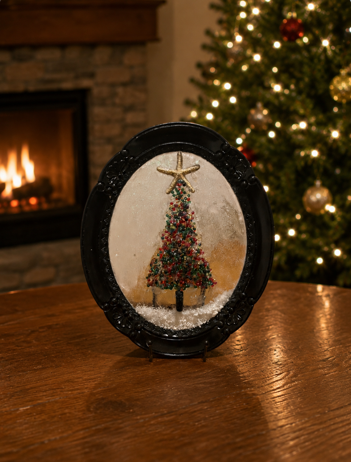 A decorative winter scene with a small Christmas tree inside a glass snow globe with a star topper, placed on a wooden table. In the background, there is a lit fireplace and a Christmas tree decorated with lights and ornaments.