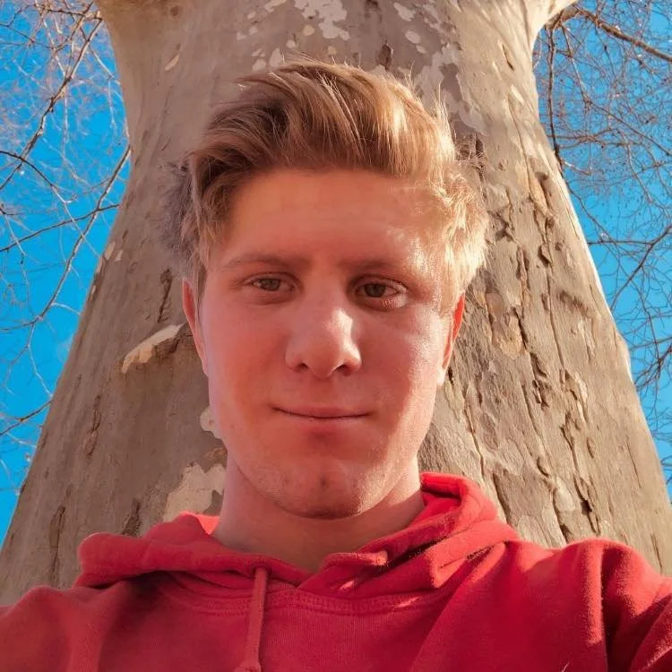Young man with light brown hair wearing a red hoodie standing in front of a tree with peeling bark and a clear blue sky.