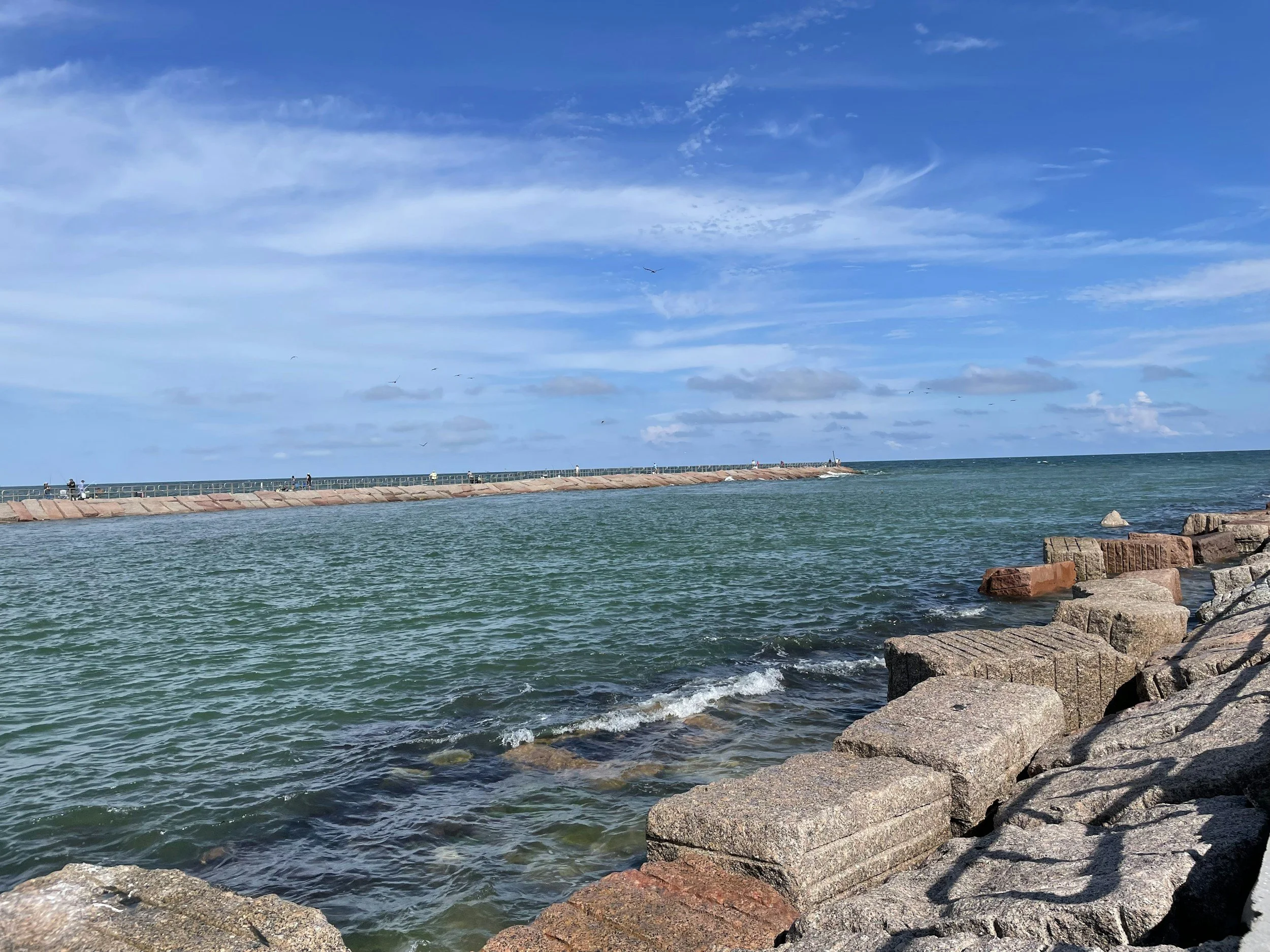 View of a breakout from a rocky shoreline into the ocean with a long pier or jetty extending into the water, under a partly cloudy blue sky with a few seagulls flying overhead.