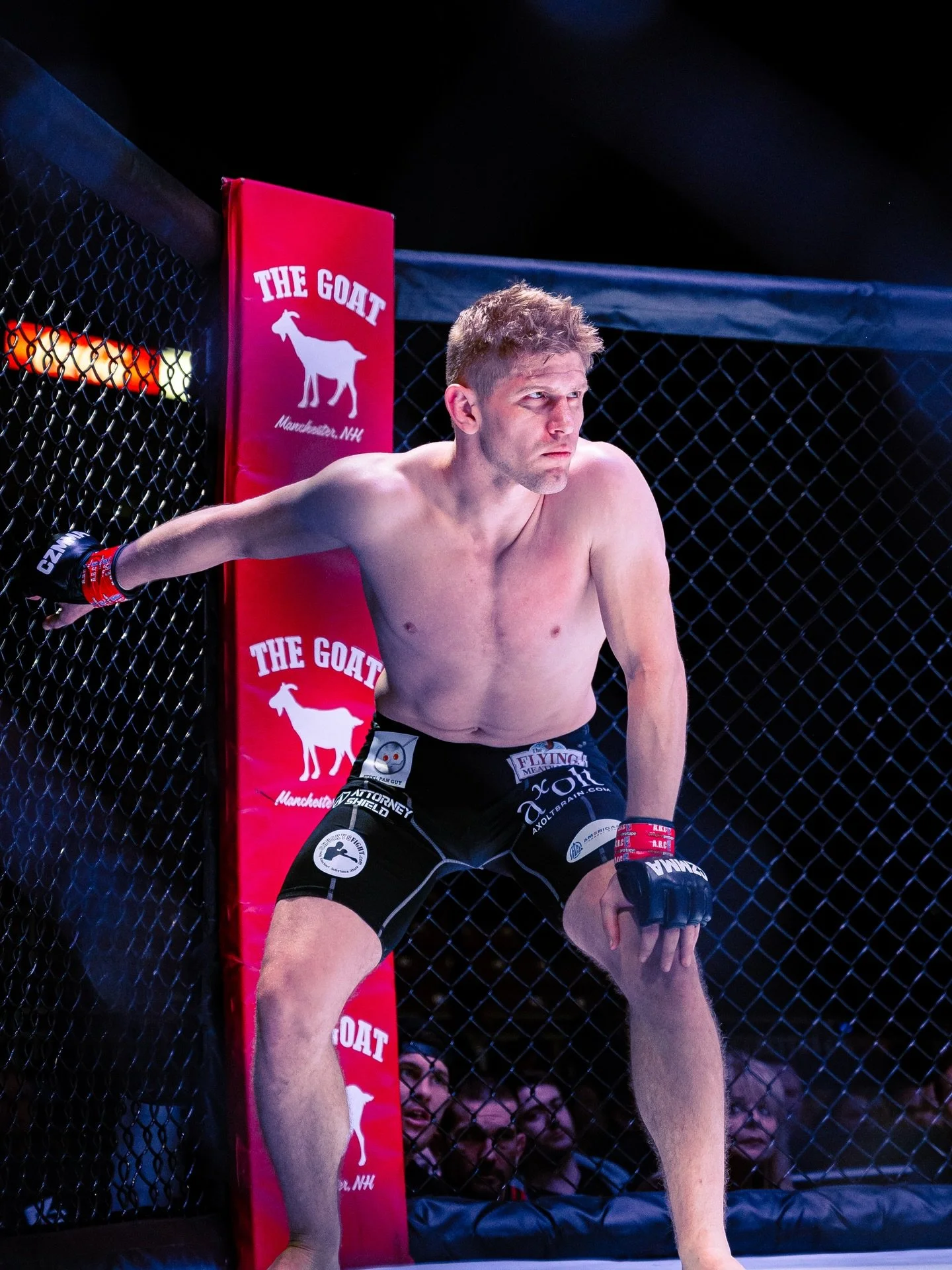Mixed martial arts fighter in black shorts and red gloves, leaning against the cage during a match.