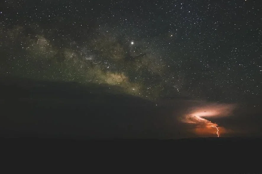 Night sky with the Milky Way galaxy and a lightning bolt in the distance.