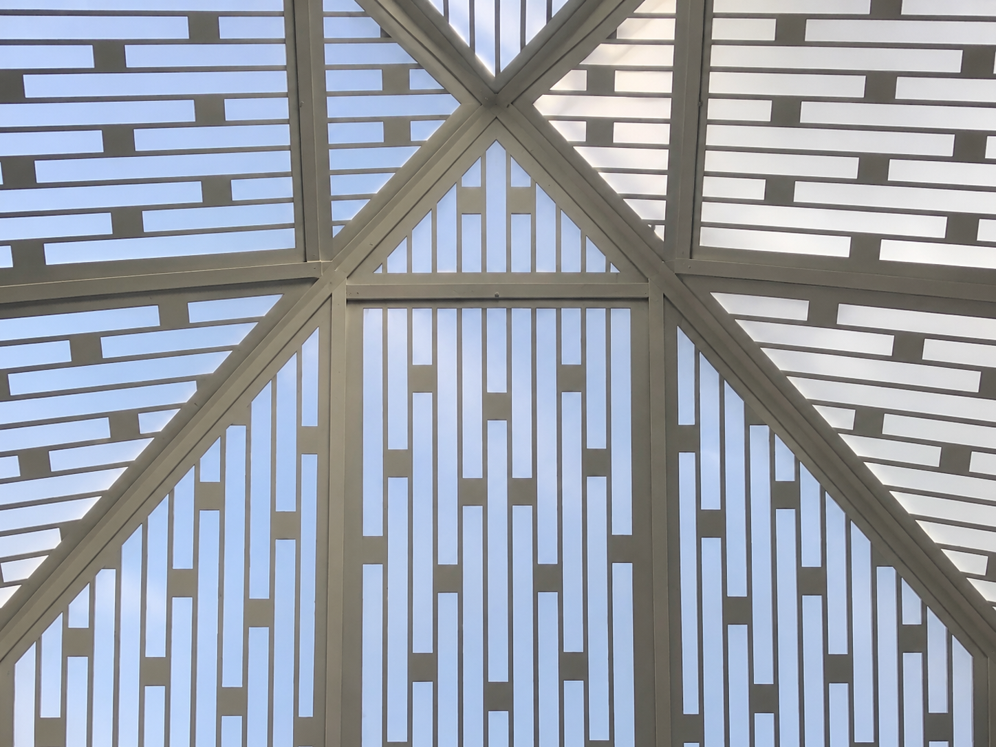 Looking up at the ceiling of a modern building with geometric metal framework and a translucent roof, allowing daylight to pass through.