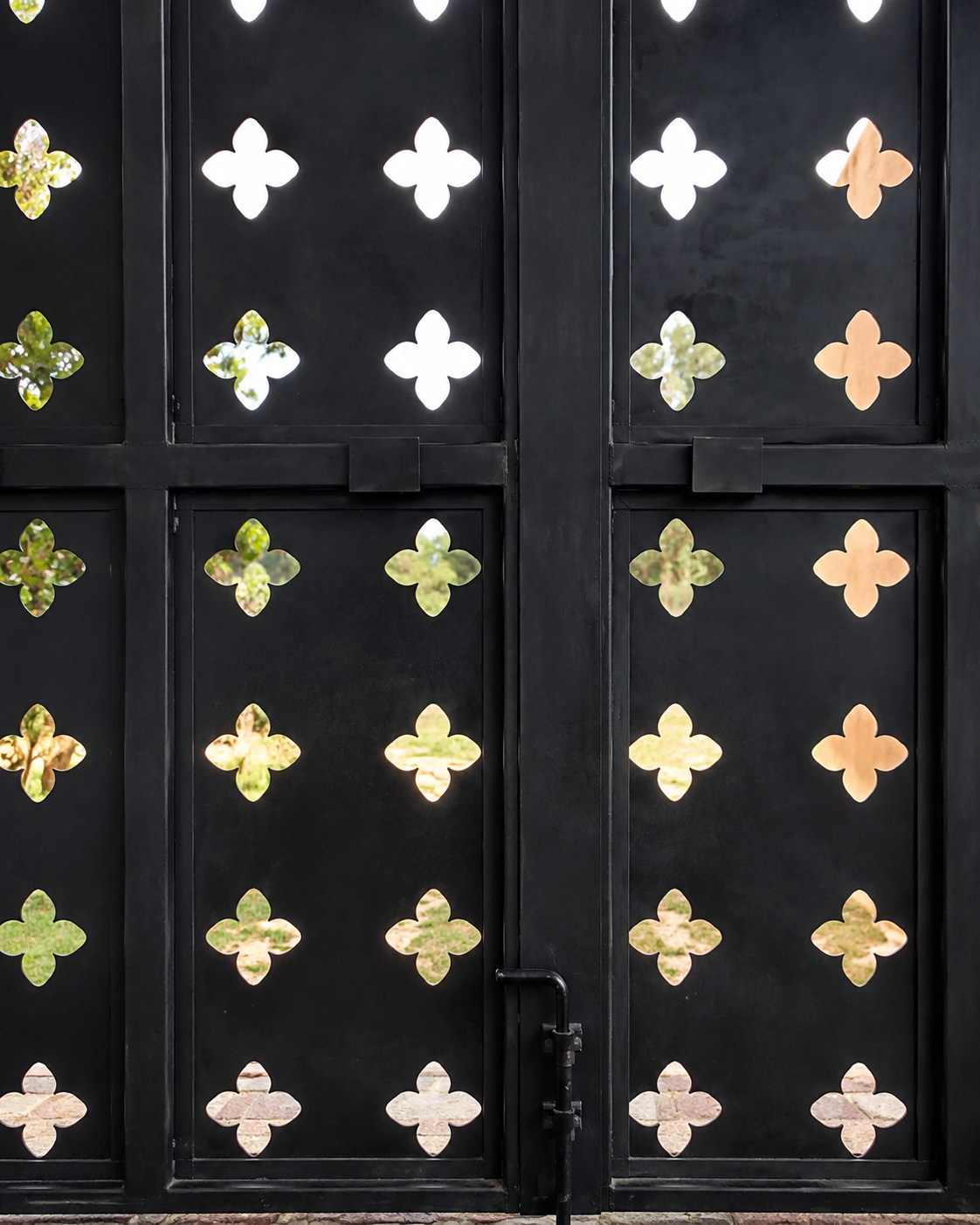 A black metal gate with a pattern of four-petaled flower-shaped cutouts, through which green trees and grass are visible.