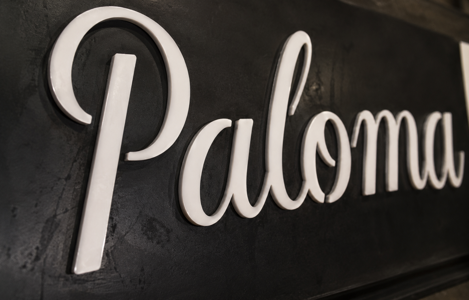 Close-up of white cursive letters spelling 'Paloma' mounted on a dark textured wall.