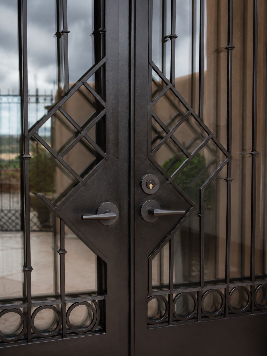 Black metal gate with geometric patterns and glass panels, handles, and a lock, reflected in nearby glass door or window.