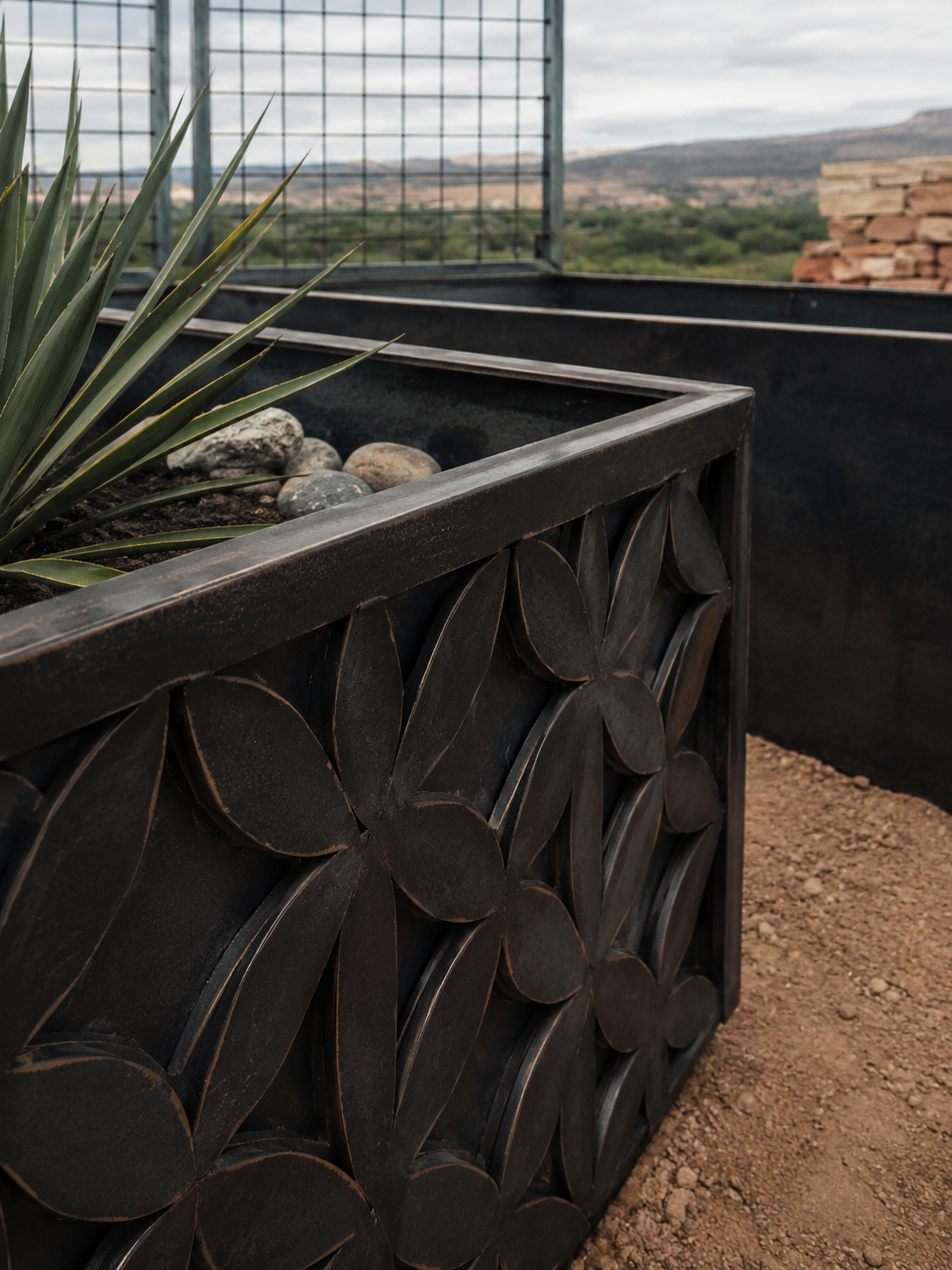 Close-up of a decorative metal planter box with a desert plant and rocks, set outdoors with a mountainous landscape and a cloudy sky in the background.