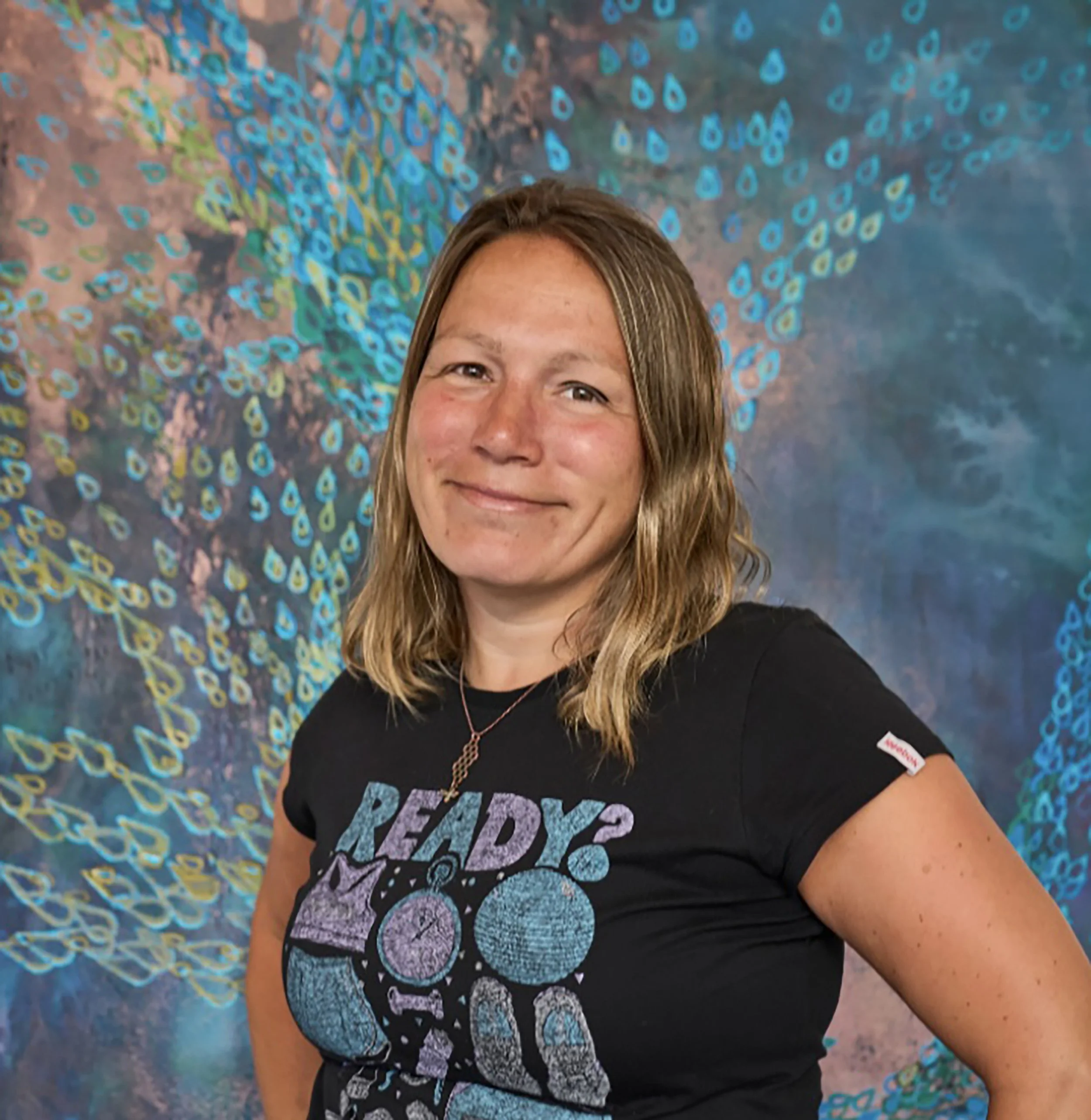 A woman with long, blonde hair smiling in front of a colorful mural of peacock feathers.