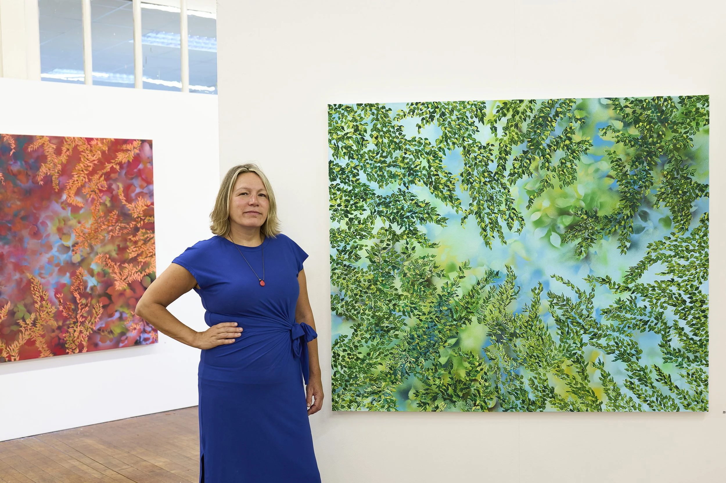 A woman in a blue dress standing in an art gallery next to large colorful photographs of green foliage and plants.