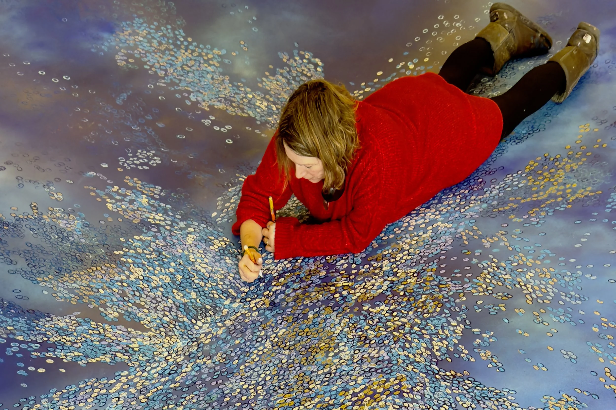 A woman in a red sweater lying on the floor surrounded by a scattered pattern of shiny multicolored coins, as if she is resting or looking at the coins.