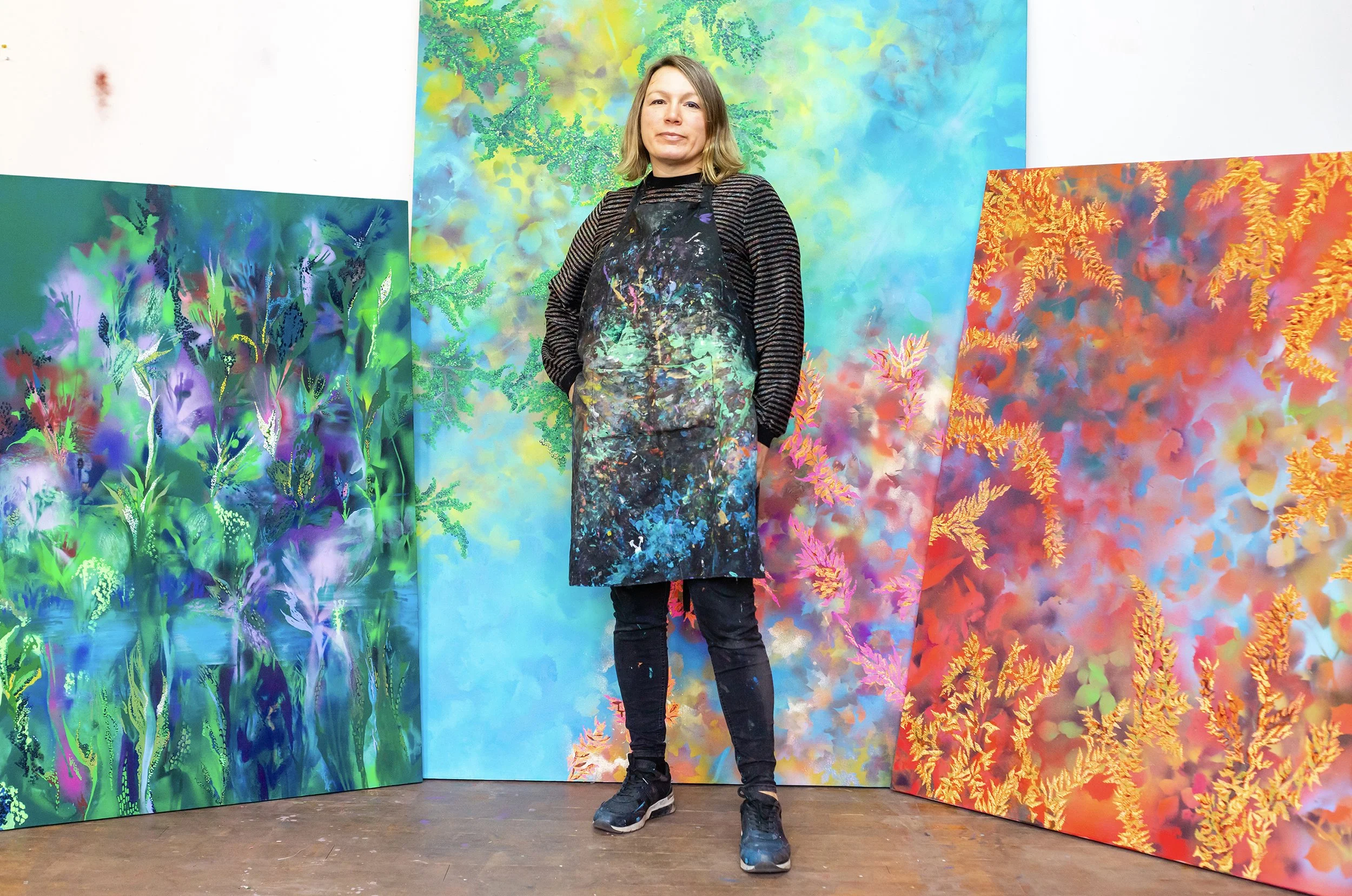 An artist standing in front of abstract colorful paintings with floral and nature-inspired designs at an art gallery or studio.