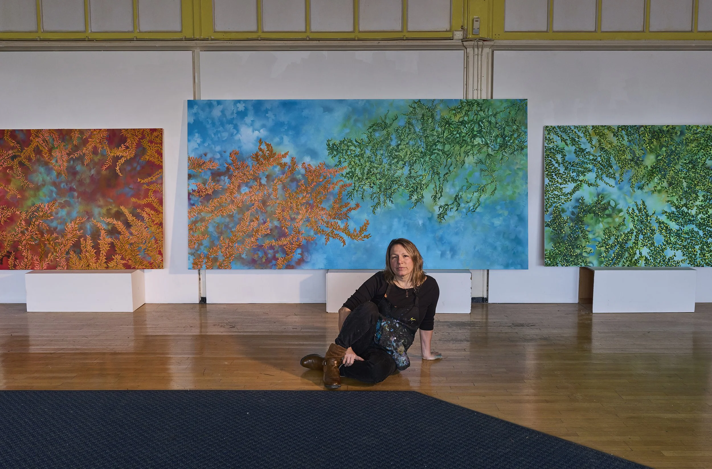 A woman sitting on the floor in front of three large colorful paintings with abstract, coral-like patterns. She is wearing a black top, paint splattered pants, and brown boots, and she is leaning against the wall in an art gallery.