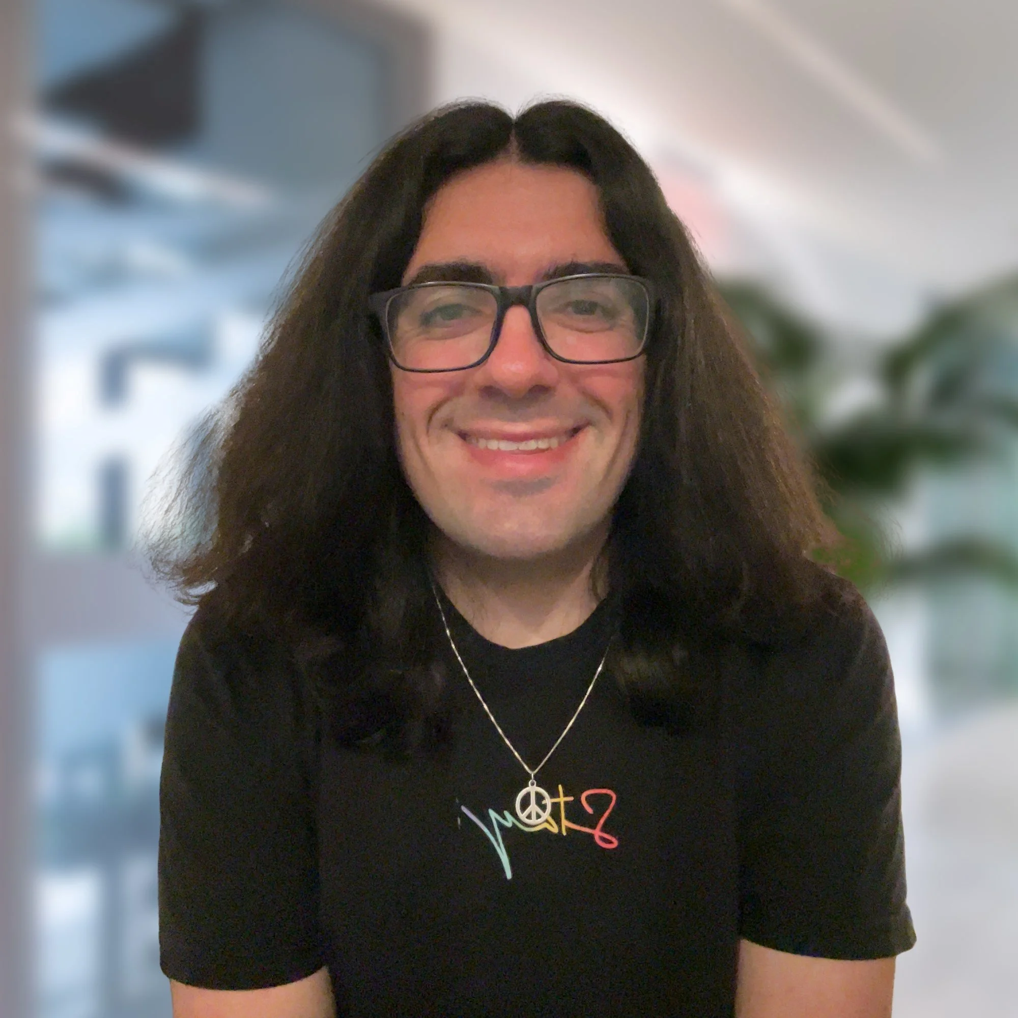 Close-up of a smiling woman with long dark hair and glasses, wearing a black T-shirt with colorful lettering and a peace sign necklace.