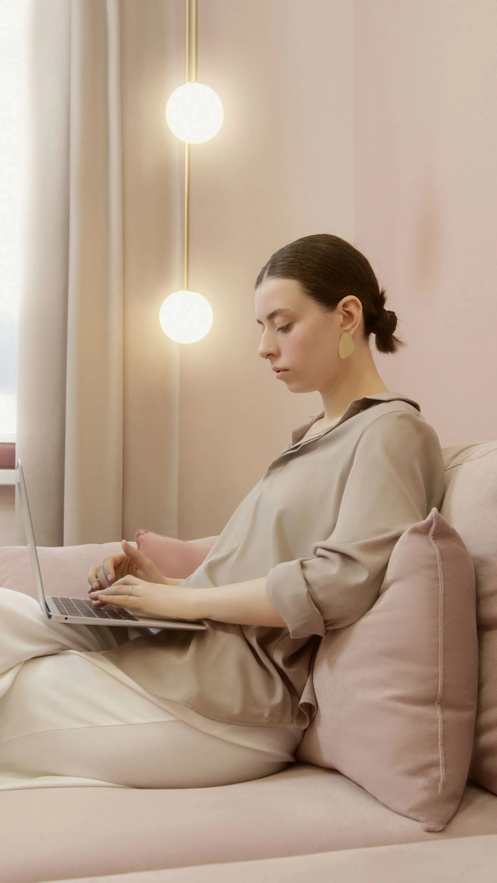 A woman with dark hair in a low bun, wearing beige clothing and gold earrings, sitting on a pink couch, working on a laptop.