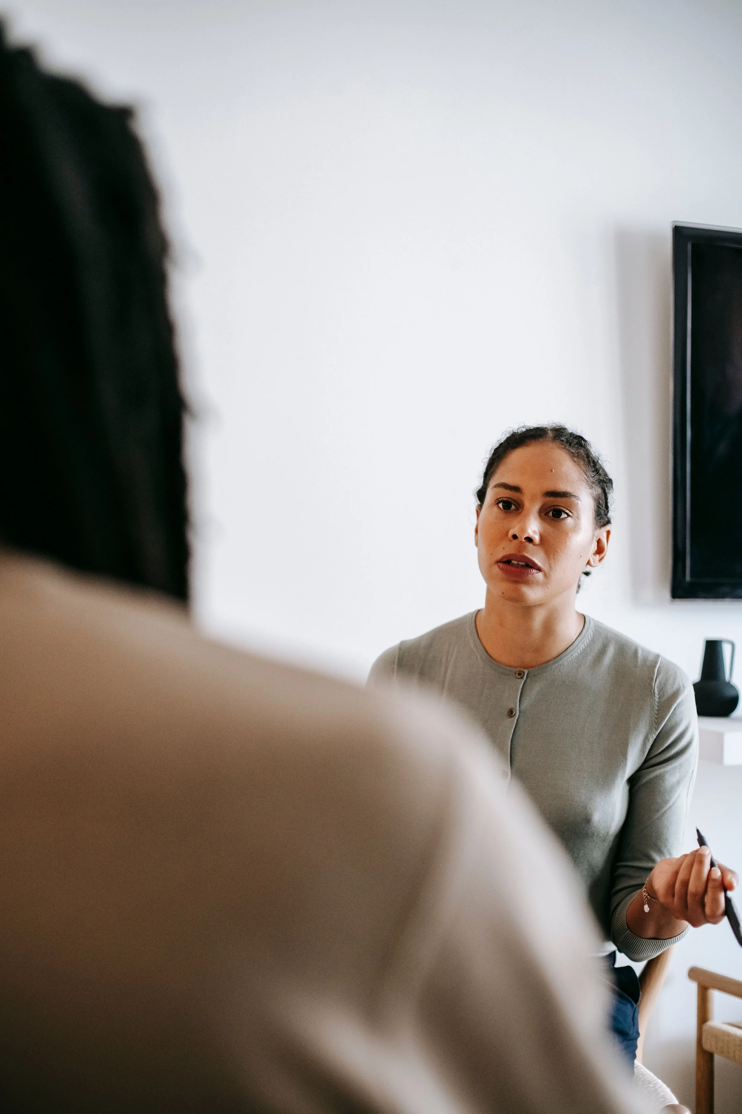 A woman with light brown hair styled in braids talking to a man who is mostly out of frame. The woman is wearing a light gray top with button details and holding a pen. They are in a room with a white wall and a TV or monitor visible in the background.