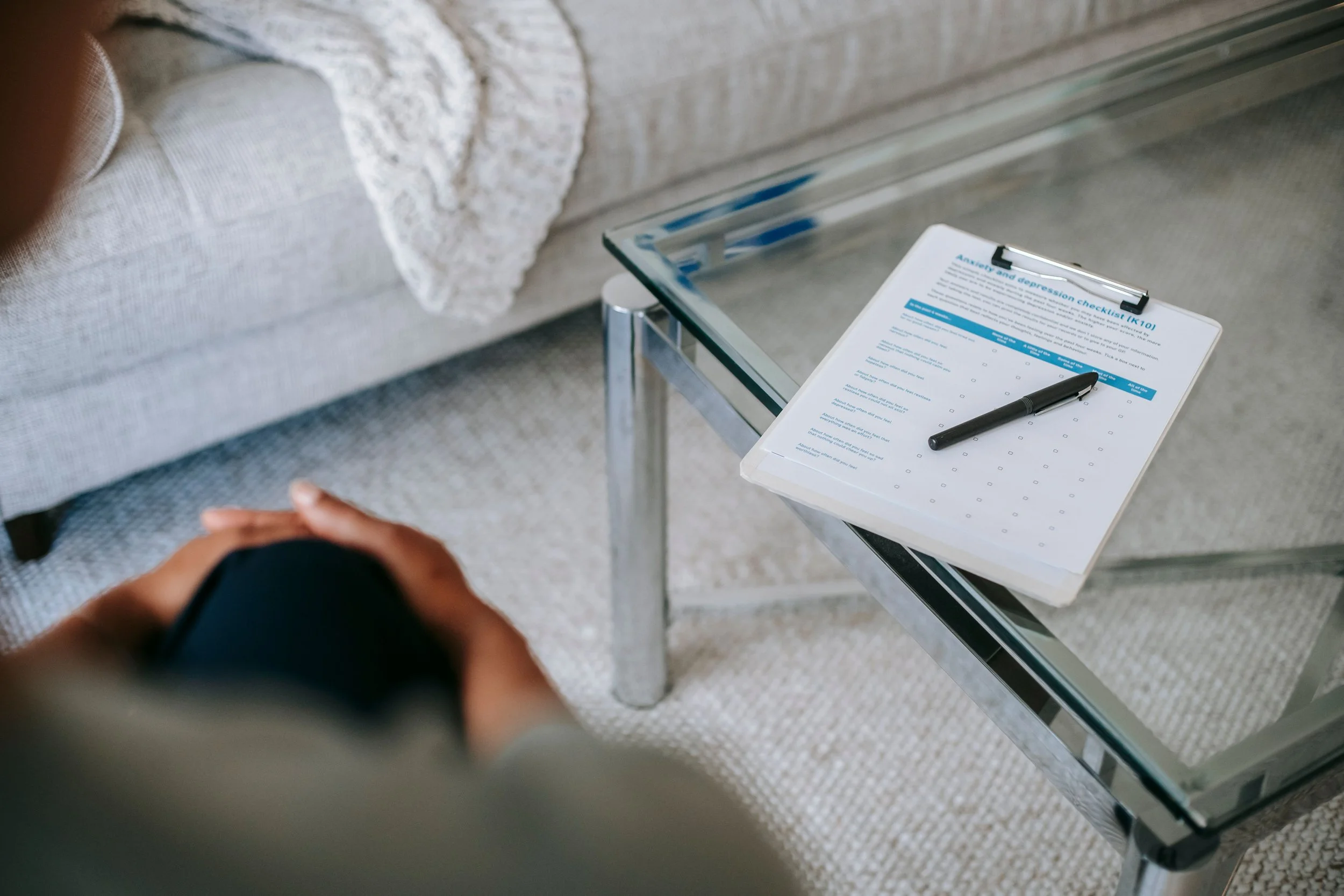 A person is seated with their hands clasped, looking at a glass table holding a checklist titled 'Anxiety and depression checklist (K10)'. A black marker pen is resting on the paper. In the background, there is a light-colored sofa with a lace throw and a beige carpeted floor.