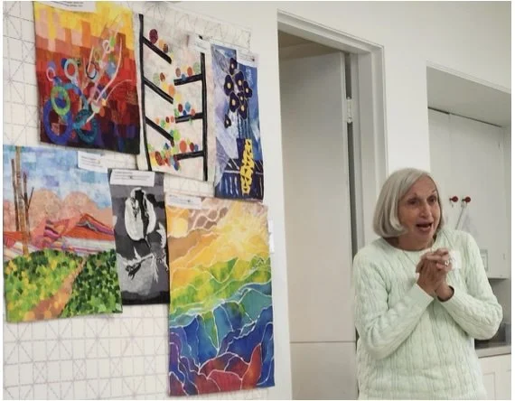An older woman with gray hair and a light green sweater is standing in front of a white wall with colorful abstract paintings hanging on it. She appears to be excited or happy, with her hands clasped together.