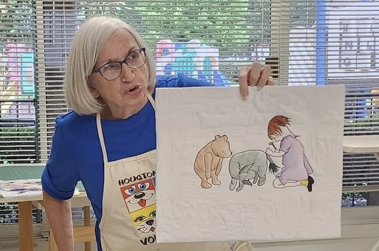 Older woman with glasses and gray hair holding a drawing of a girl and animals, inside a room with windows and blinds