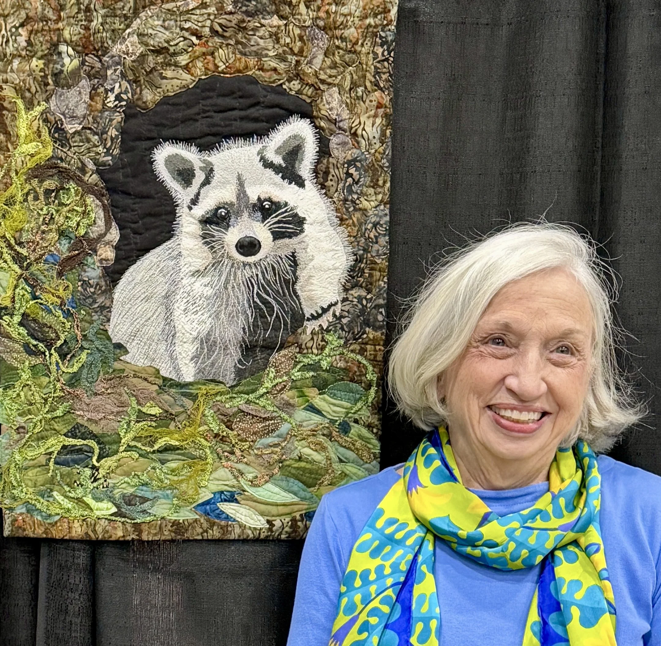Close-up of a woman with white hair smiling in front of a dark wall and a quilted art piece featuring a raccoon surrounded by abstract fabric and decorative moss.