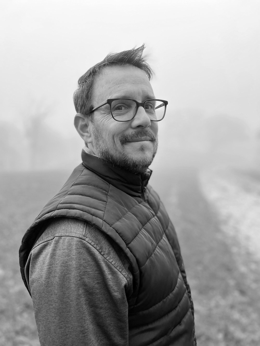 A black and white portrait of a man with glasses and a beard, smiling slightly, wearing a quilted vest over a long-sleeve shirt, standing outdoors with a foggy background.