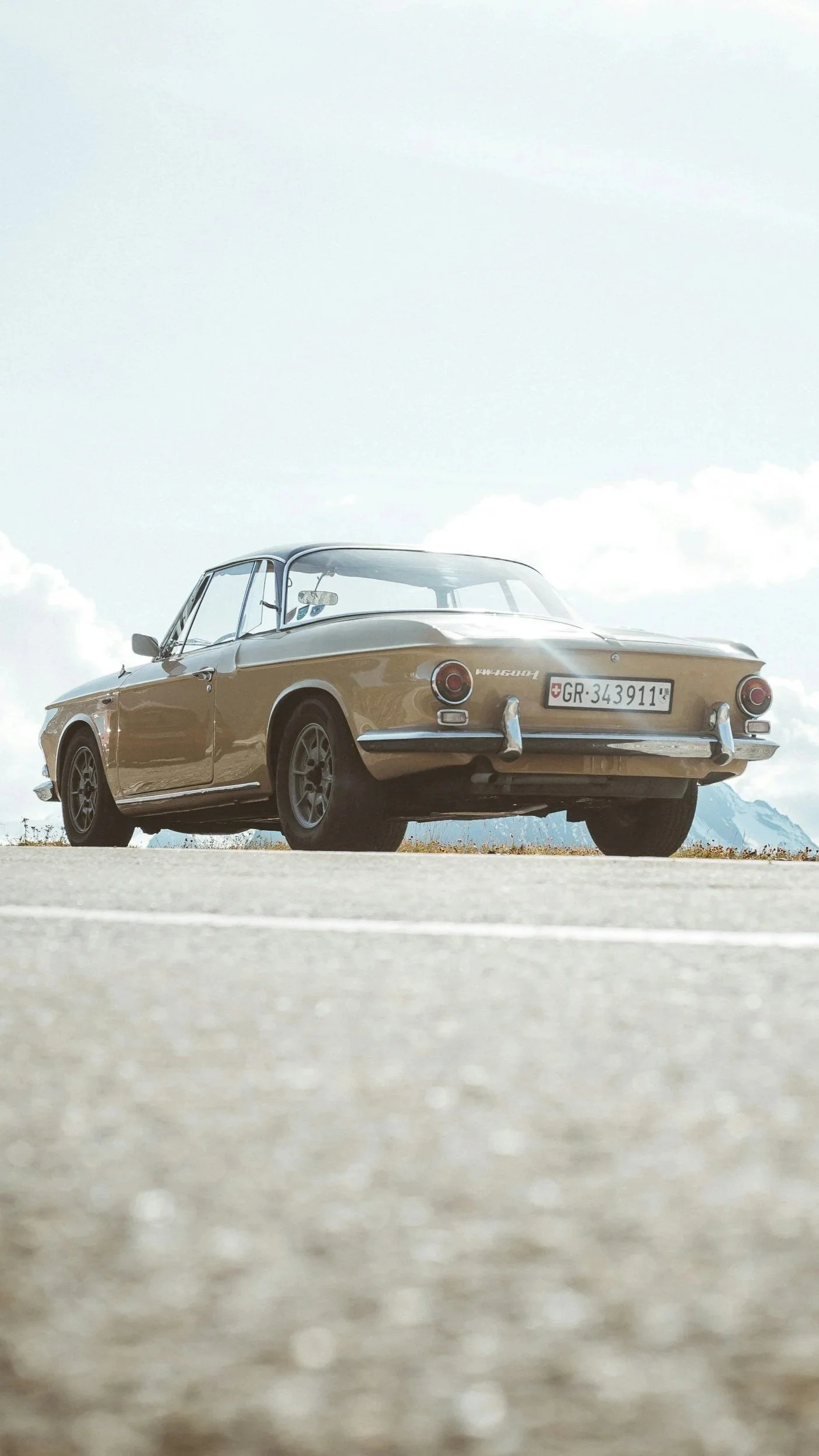 A vintage beige car parked on an open road with mountains in the background and a cloudy sky.