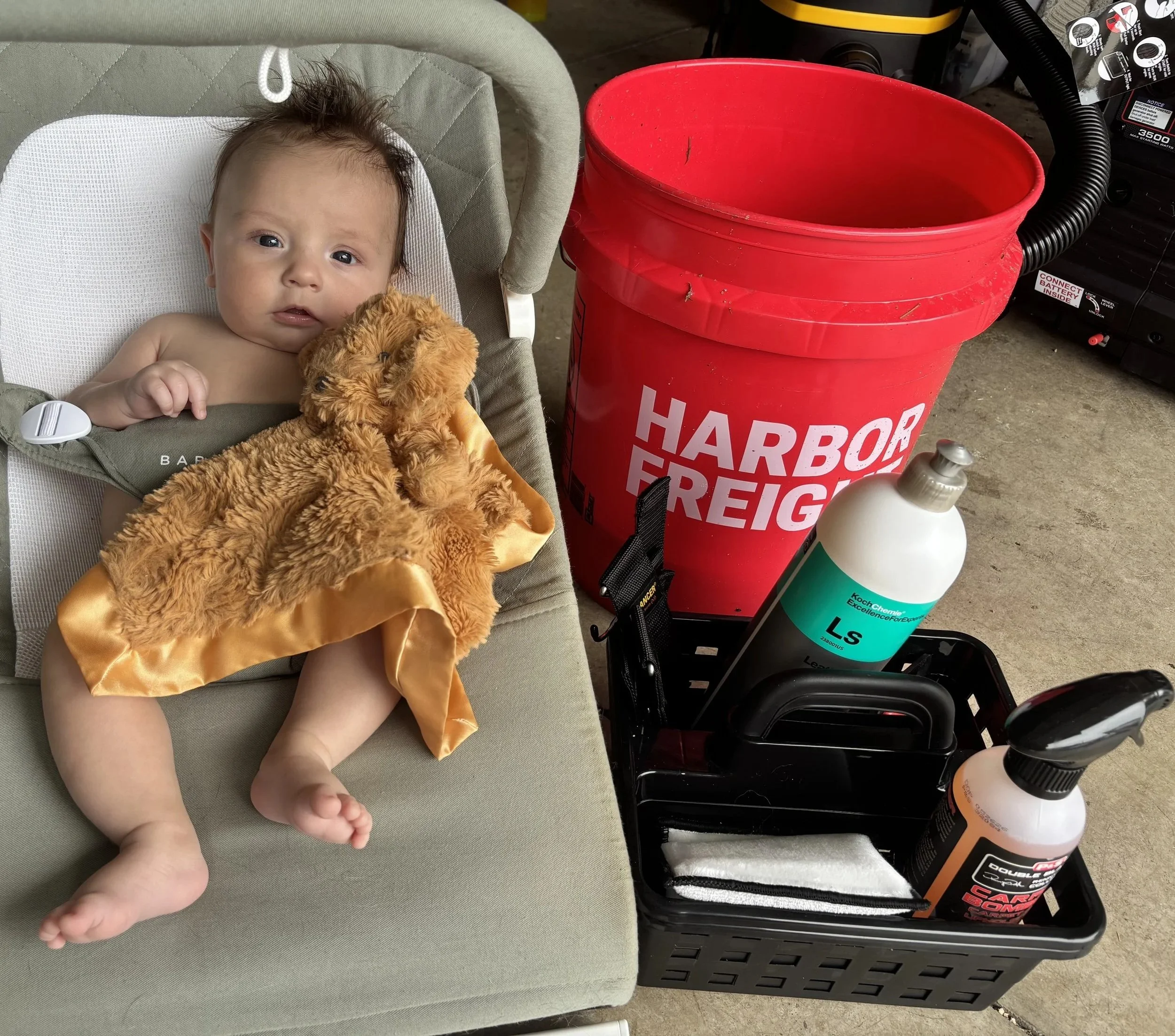A baby sitting in a car seat holding a stuffed animal with a red bucket and cleaning supplies nearby.