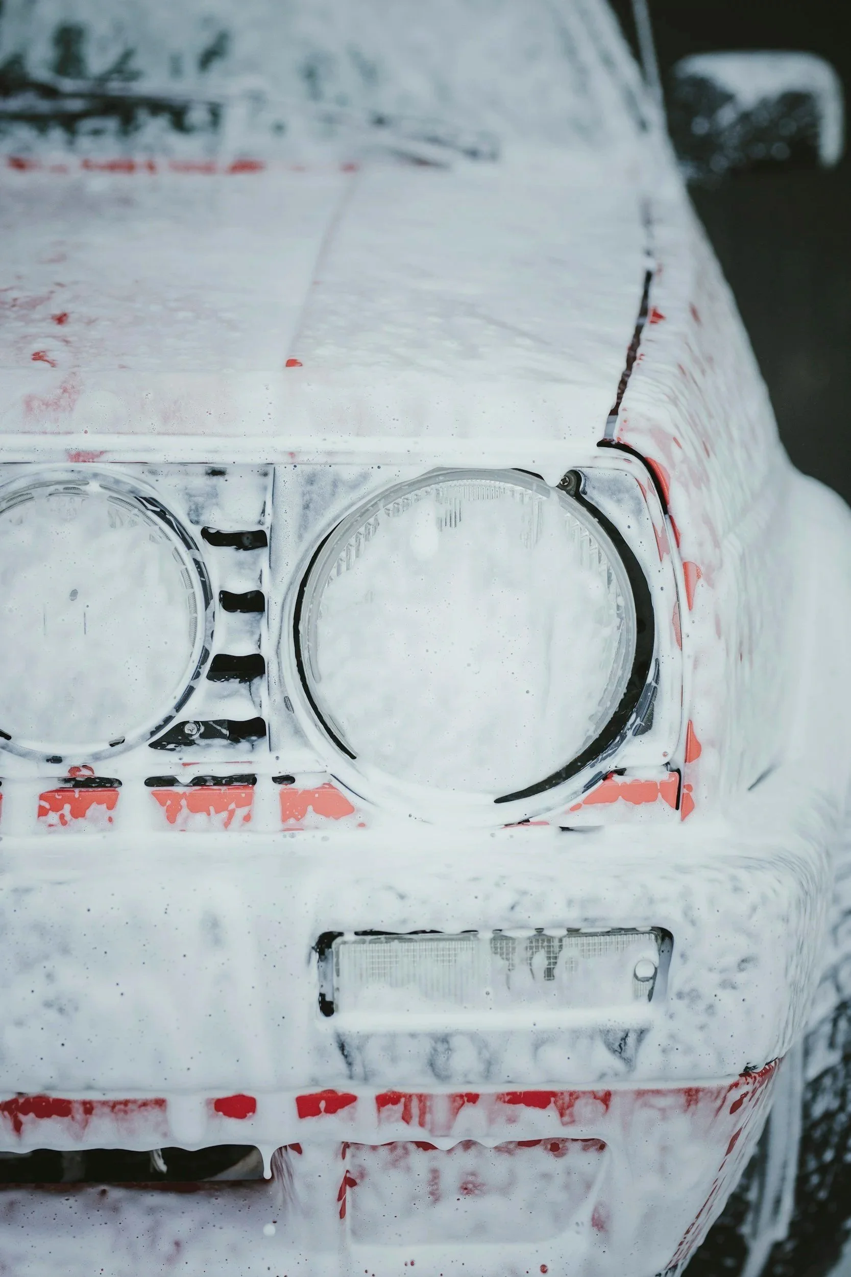 Front of a foamy soapy vehicle with visible headlight outlines, parts of the bumper, and a license plate, all partially obscured by soap.