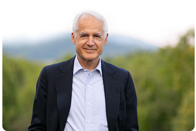 Hunter Lewis wearing a dark suit jacket and a light blue dress shirt outdoors, with blurred greenery and mountains in the background.