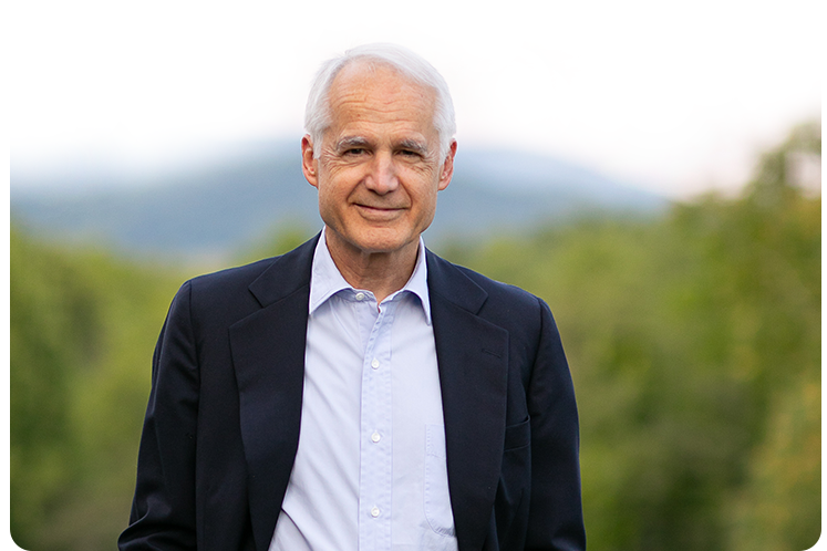 Hunter Lewis dressed in a dark blazer and light shirt standing outdoors with green trees and mountains in the background.