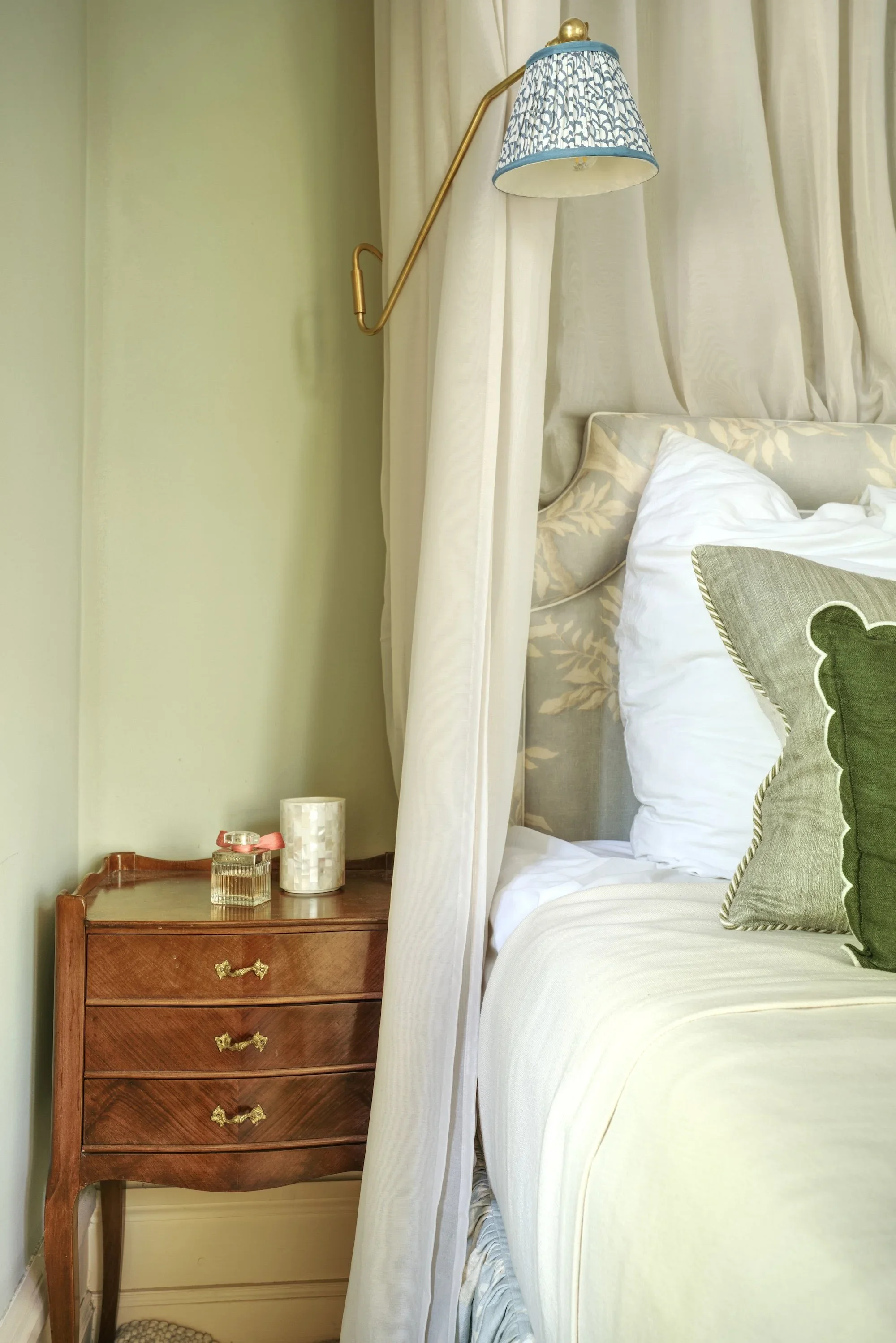 Headboard in Lewis & Wood Doves fabric against Farrow & Ball French Gray walls, beside an antique chest of drawers and a brass wall light