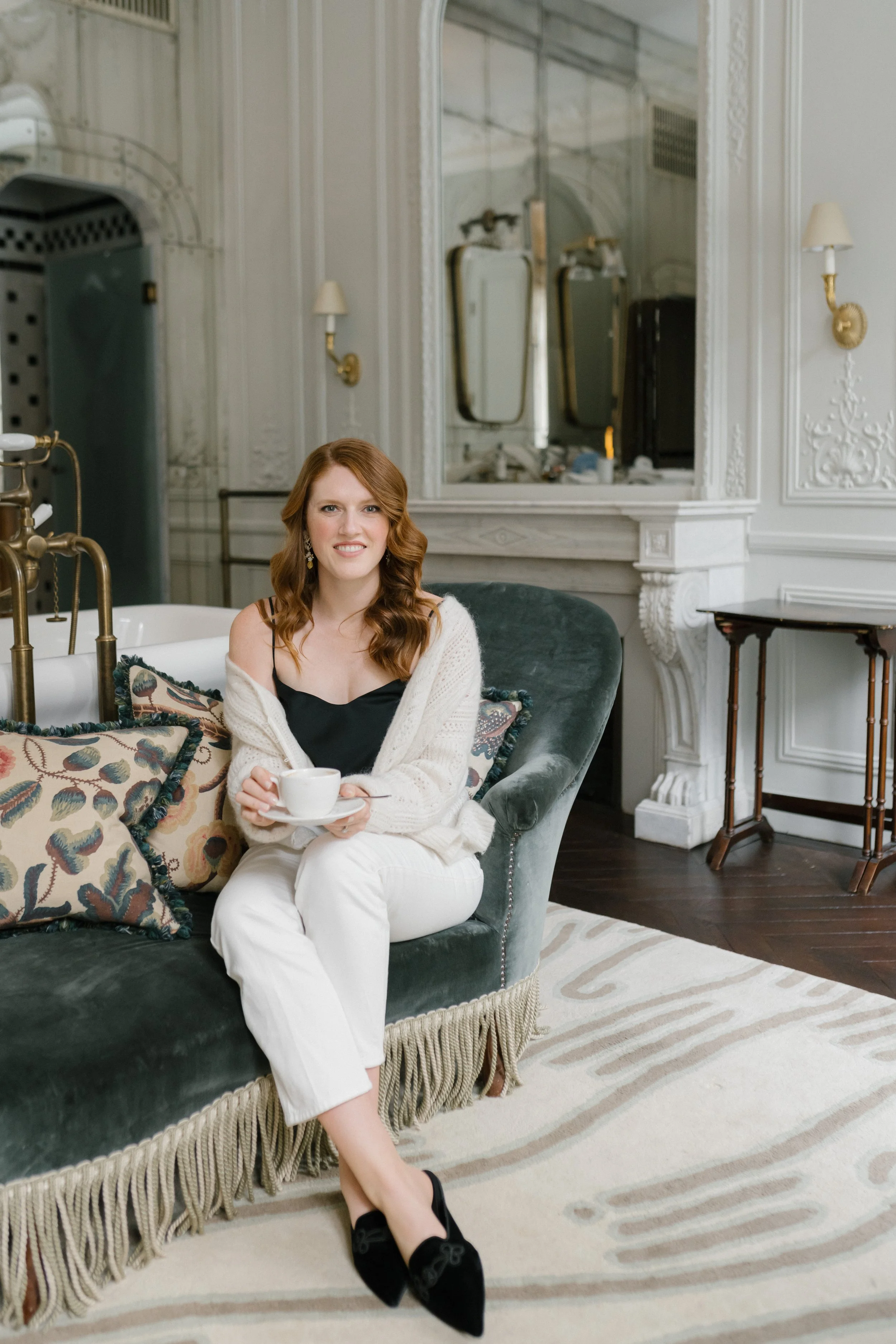 A woman with curly red hair sitting on a dark green velvet sofa, holding a white teacup and saucer, smiling at the camera in a luxury room with white ornate walls, wall sconces, a large mirror, and a patterned rug.