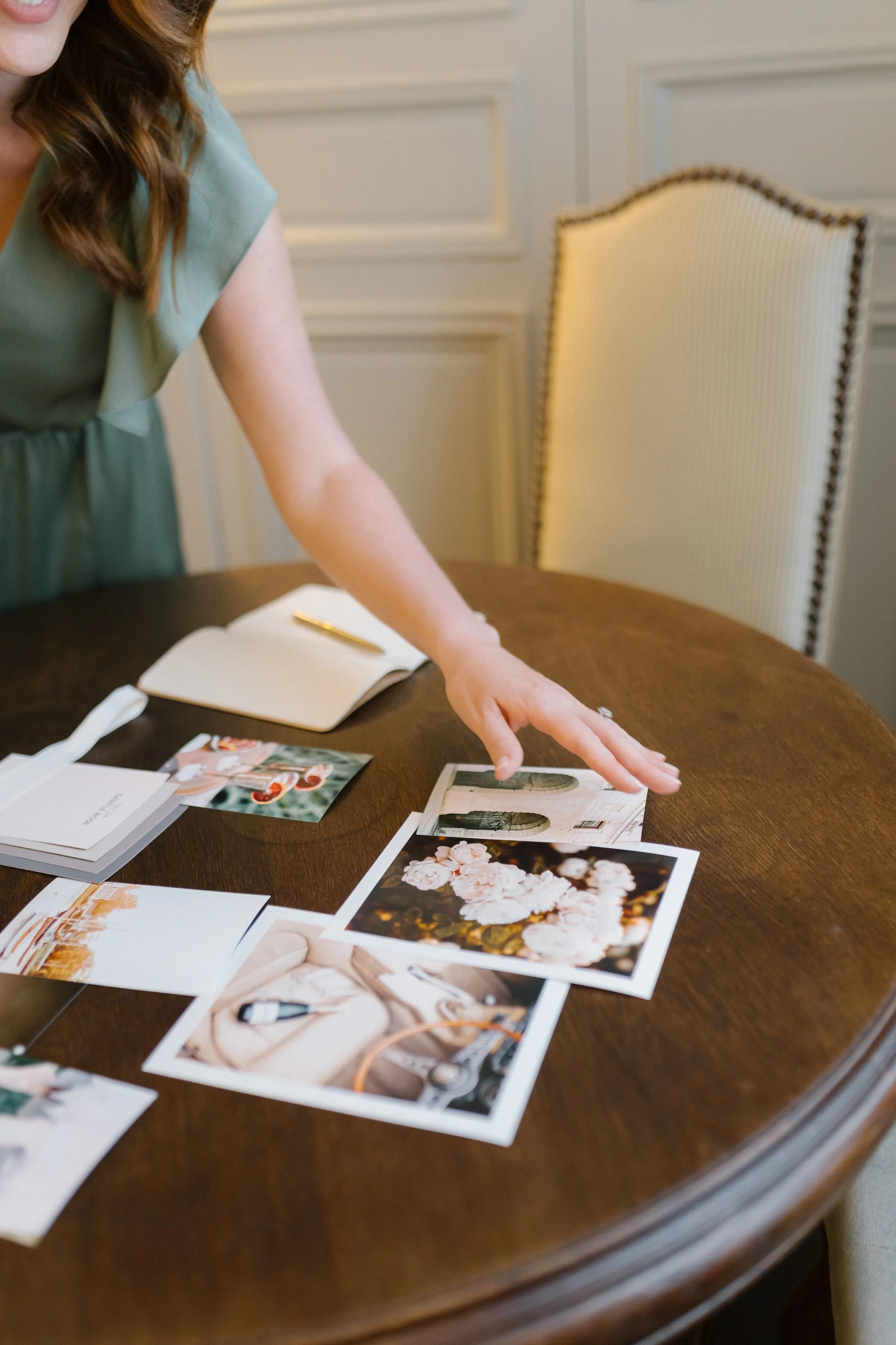 Close-up of a woman with reddish hair, in a light green dress, pointing at photographs on a wooden table in an elegant room with beige paneled walls and a high-backed ivory chair.