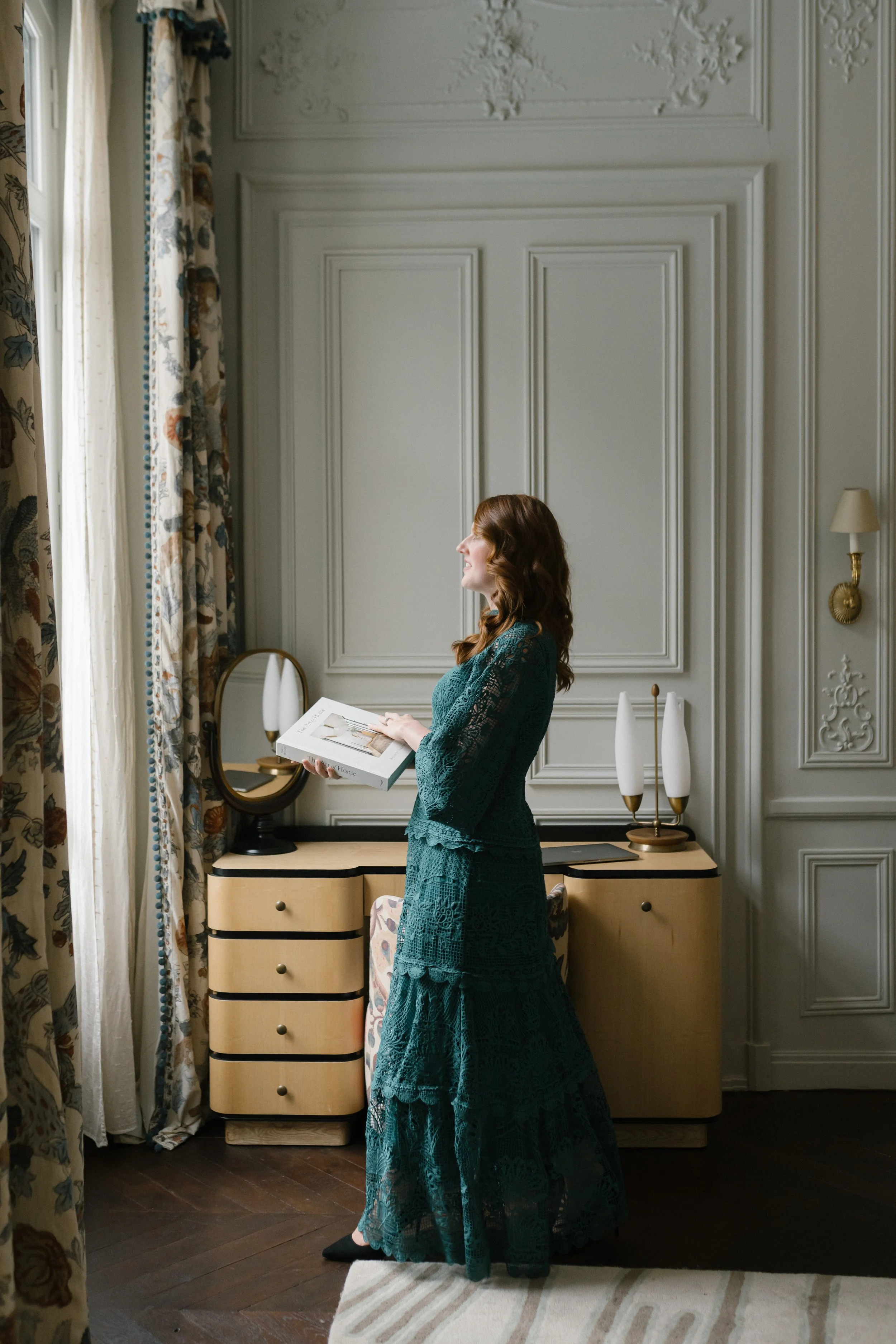 A woman with long red hair in a teal lace dress is standing indoors, smiling, while holding a white box with what appears to be documents or papers inside. She is positioned in front of a white wall with decorative molding, next to a dresser with white and black accents, topped with two white lamps and a small mirror. There is a beige and white area rug and floral curtains on the window to her left.