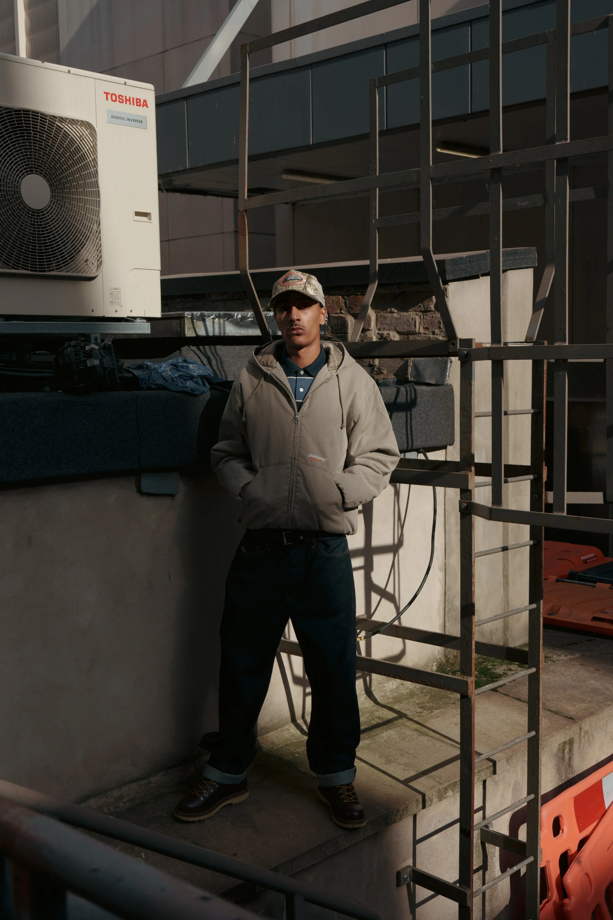 A young man stands outside near a building, wearing a beige hoodie, dark jeans, black boots, and a baseball cap. He is positioned in front of a metal scaffolding and a section of brick wall, with construction materials and equipment nearby.