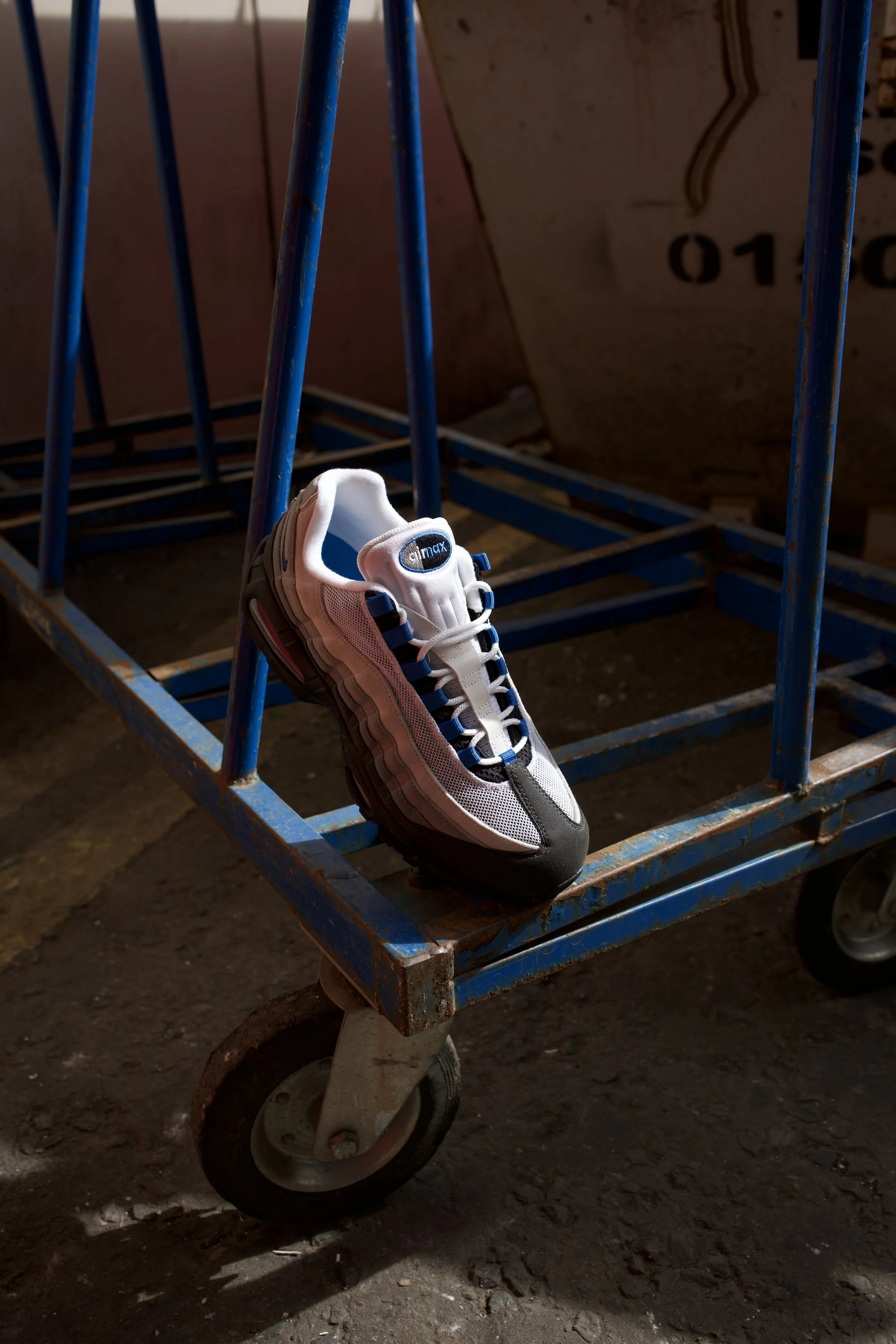 A men's athletic shoe resting on a blue metal cart with caster wheel in a dimly lit space with a concrete floor and corrugated background.