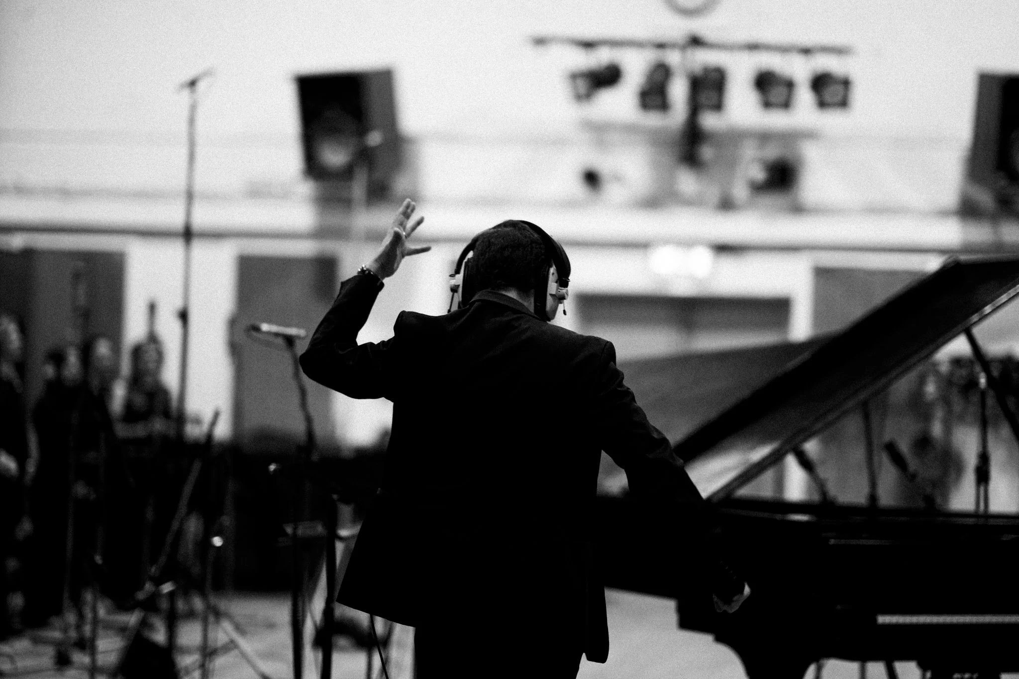 A person in dark clothing and headphones is conducting a piano in a music studio. The person is facing the piano and raising one hand, likely conducting or playing. The studio has various electronic equipment and soundproofing panels.